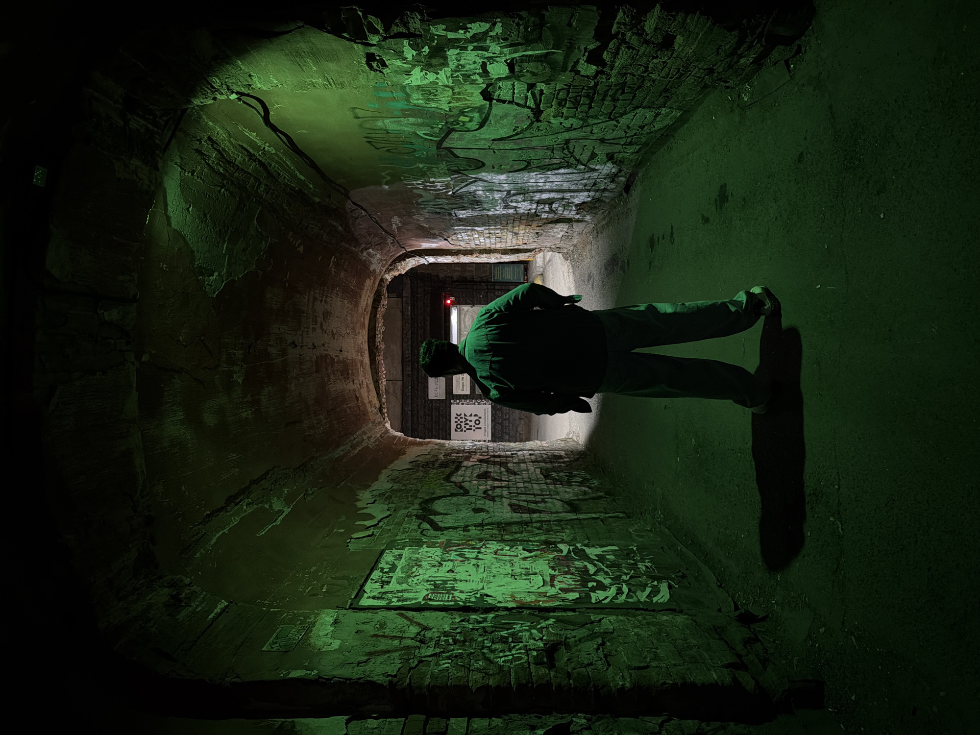 A low-angle, indoor shot depicts a tunnel illuminated by green light. The tunnel has brick walls and ceiling with visible damage and graffiti. A man stands in the center of the tunnel, facing away from the camera, wearing a dark jacket and light-colored pants. His shadow stretches onto the floor in front of him. At the far end of the tunnel, there is an entrance visible with posters on the walls. Based on the tags the location is likely in Krasnoyarsk, Russia.