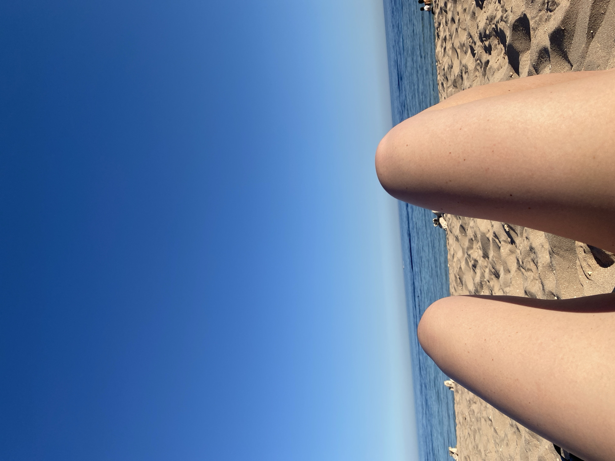 The image depicts a view from the perspective of someone lying on a sandy beach. The foreground is dominated by the thighs and knees of an individual, casting shadows on the sand. The beach is composed of fine, light-colored sand with visible footprints and small indentations. Beyond the sand, a calm blue sea stretches to the horizon, meeting a clear, bright blue sky. In the distance, along the coastline, several small figures of people can be seen, some standing and some possibly sitting. The overall scene suggests a sunny day at a beach resort, likely in a location like Sharm El Sheikh, Egypt, known for its coastal beauty and clear skies.