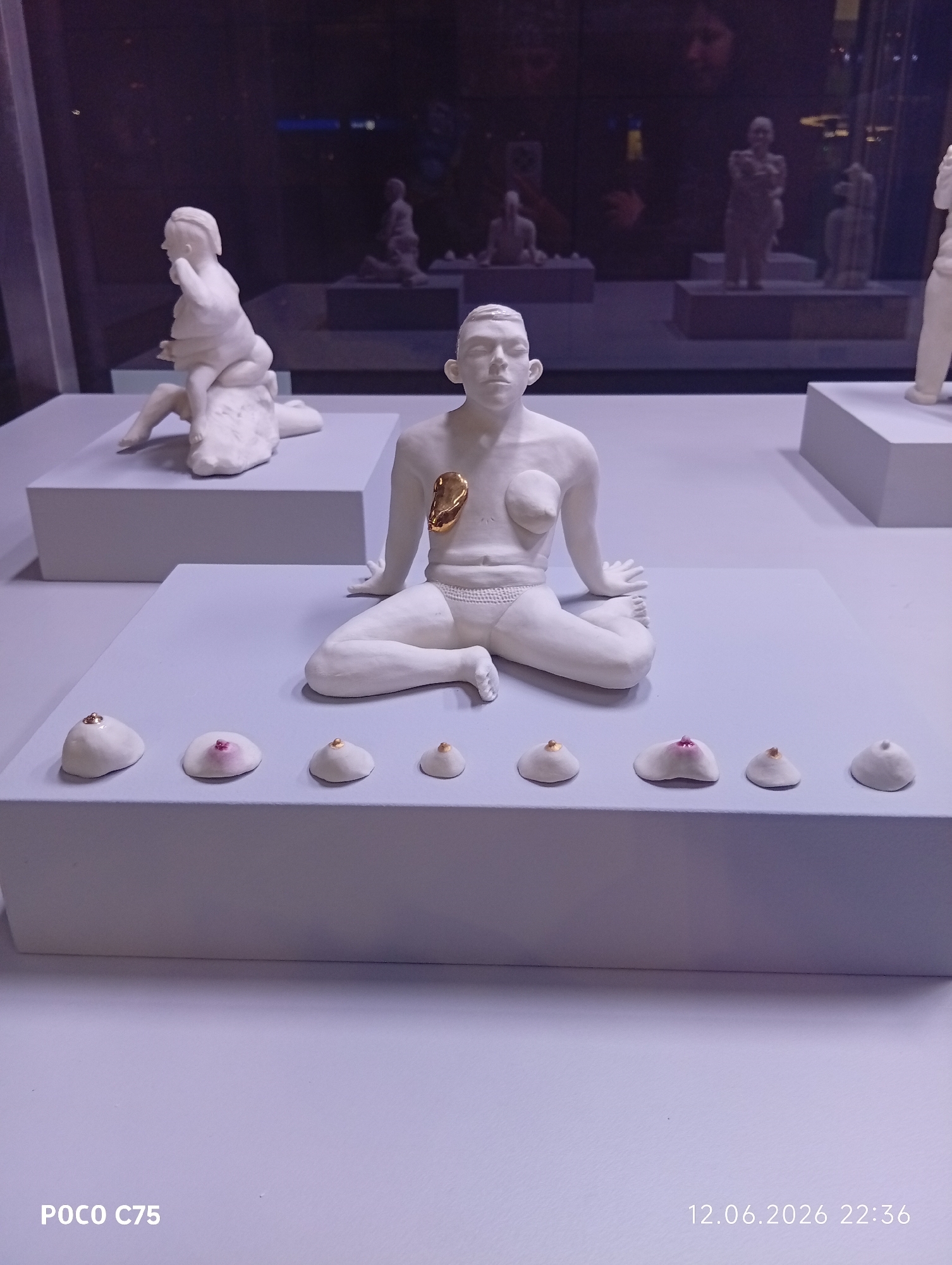 The image shows a display of white ceramic sculptures, likely in a museum or gallery setting. In the foreground, a large sculpture of a seated nude figure is the central focus. This figure has a prominent gold-colored adornment over its chest. In front of this figure, on a lower white platform, are several smaller, breast-shaped ceramic objects, each with a small gemstone or decorative element on top. Behind the central figure, further white sculptures are visible on raised platforms, including one of a figure in a seated, intertwined pose. The background appears to be a dimly lit display case with more sculptures, and reflections of people can be seen on the glass. The lower right corner of the image displays a timestamp: "12.06.2026 22:36". The lower left corner shows "POCO C75", indicating the device used to capture the image. The scene is likely an art installation. Without additional information, the specific location in Wrocław, Poland cannot be determined from the image alone.