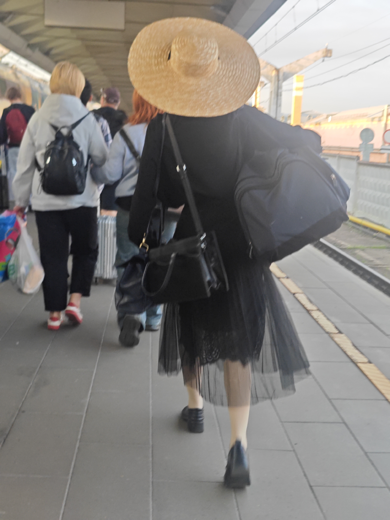 The image depicts a view from behind of several people walking on a train platform. The foreground is dominated by a woman wearing a wide-brimmed straw hat, a black coat, and a sheer black skirt, carrying a large blue duffel bag over her shoulder and a black handbag. To her left, a woman in a grey jacket and black pants walks with a black backpack and a plastic shopping bag. Further back, other individuals are visible, including one in a light grey jacket and another in a blue shirt. The environment appears to be an outdoor train station with a tiled platform and tracks visible in the background. The lighting suggests it is daytime, possibly late afternoon, with warm light illuminating the scene. There is no specific indication of the city of Moscow, Russia within the image itself.