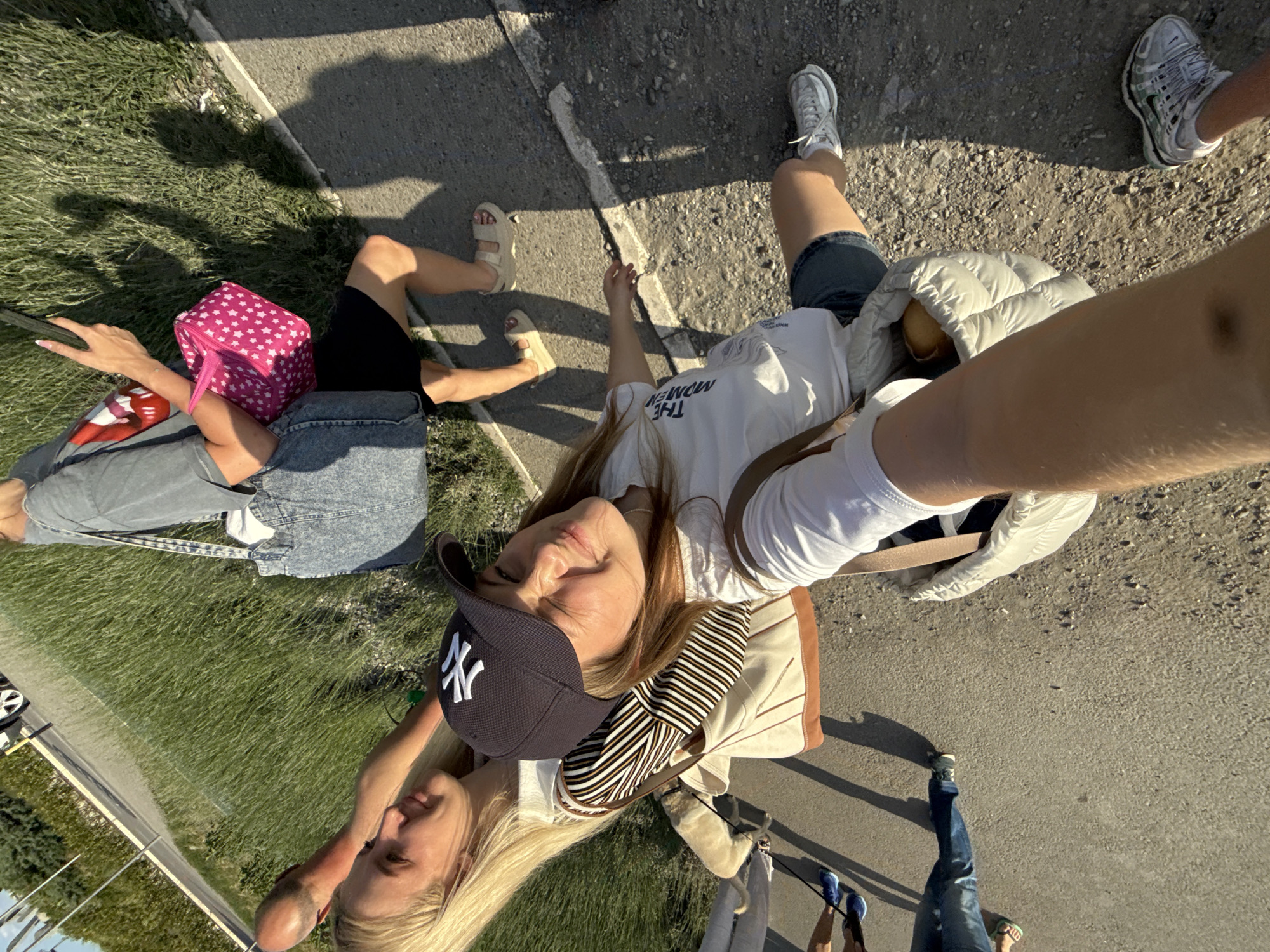 Observed from an oblique aerial perspective, the scene depicts several individuals sitting and standing adjacent to a paved surface and a grassy area. It appears to be outdoors, likely in Cukarica Urban Municipality, Serbia.
A woman in a dark New York Yankees baseball cap, white T-shirt, and jacket is seated on the edge of the pavement with another person who appears to be holding a leash. The other individual wears a black shirt, jeans, and sandals. Beside her is a pink patterned bag.
A second individual is seated on the pavement, with another wearing a grey jacket standing nearby.
The environment consists of a paved area and a grassy patch.