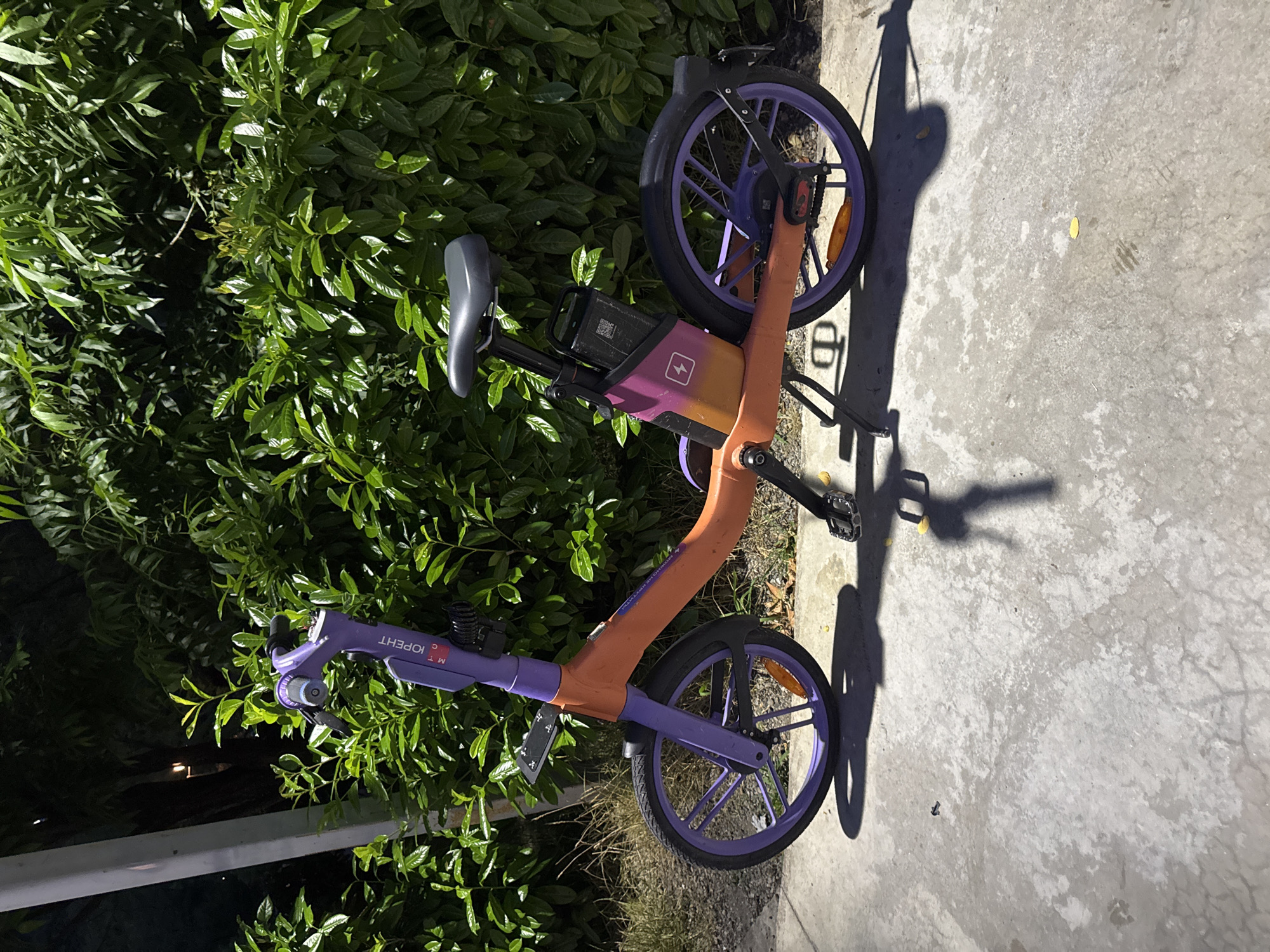 An electric scooter is parked on a concrete sidewalk in front of a dense green hedge. The scooter has an orange frame and purple wheels and handlebars. A black seat is positioned over a black battery pack. The scooter is stationary, and no people are visible. The background consists of lush green foliage, suggesting an outdoor environment. A tall, light-colored pole is partially visible to the left of the scooter. The scene appears to be in Sochi, Russia, based on the presence of Cyrillic text on the scooter's handlebar, which reads "МТС ЮRENT".