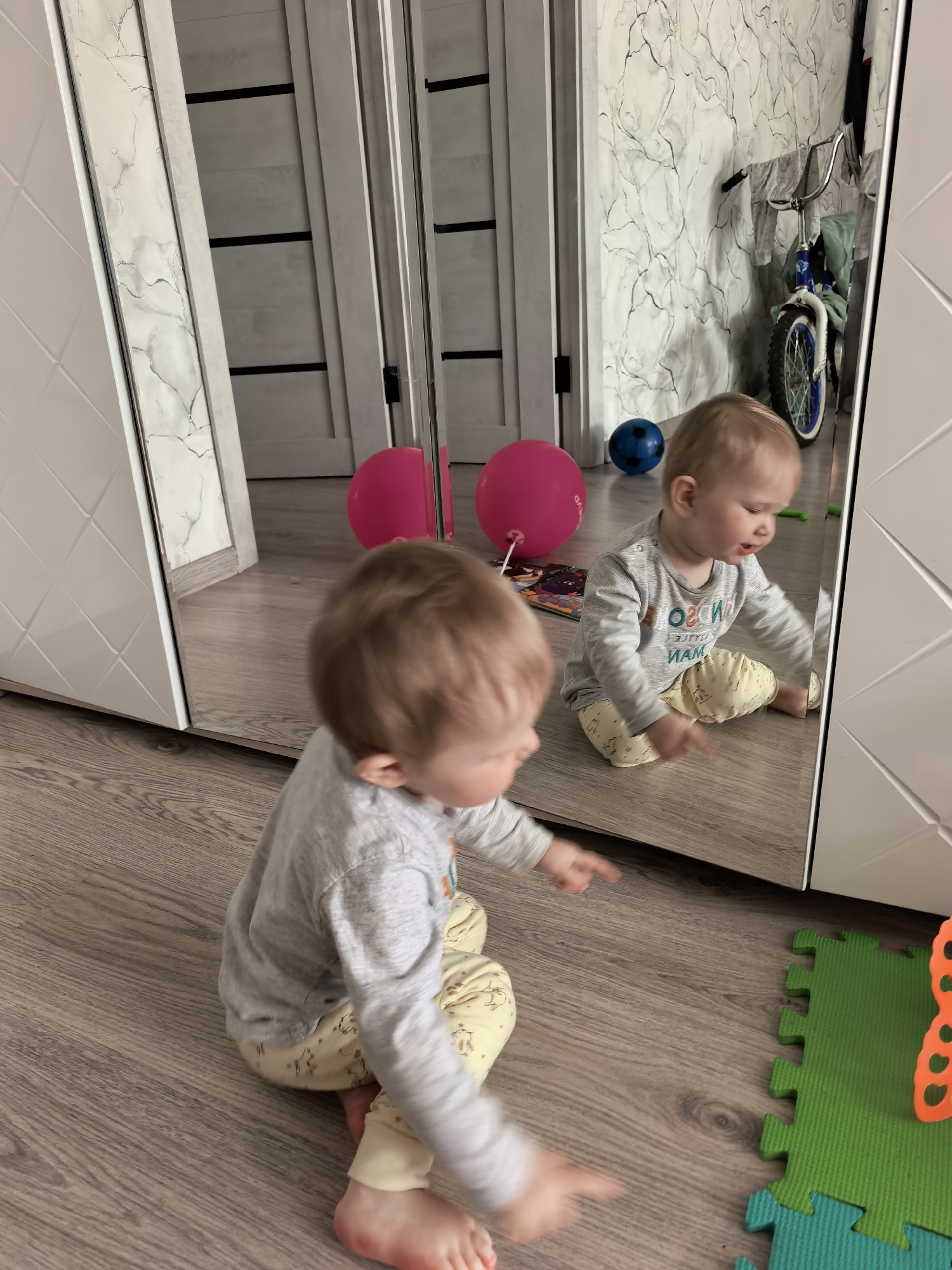 This indoor photograph, taken in Sterlitamak, Russia, features a baby or young toddler interacting with its reflection in a large, full-length mirror. The baby is seated on a light wood-patterned laminate floor, wearing a grey long-sleeved shirt with reversed text (which appears to read "LITTLE MAN") and light yellow patterned pants. The baby in the foreground is slightly out of focus, with its back partially to the viewer, and appears to be pointing or reaching towards its reflection.
The mirror itself forms the central element of the scene, reflecting the baby and parts of the room behind it. In the reflection, the baby is clearer and appears engaged, looking at itself. On the floor within the reflection, a vibrant pink balloon and a blue ball are visible, alongside a children's book or colorful toy. Further back in the reflection, a bicycle with a blue front wheel and handlebars can be seen. The wall behind the reflected objects is covered in a distinctive light-colored wallpaper with dark, swirling or marbled patterns.
The mirror is set into a piece of light-colored furniture, possibly a wardrobe or cabinet, which has a textured, possibly quilted, design on its right side. To the left of the main mirror, another section of mirrored surface with a white and grey marble-like pattern is visible, adjacent to a white wall with a subtle geometric pattern. On the floor in the foreground, to the right, green and blue interlocking foam playmat pieces are present. The scene suggests a bright, daytime setting in a child's play area or living space.