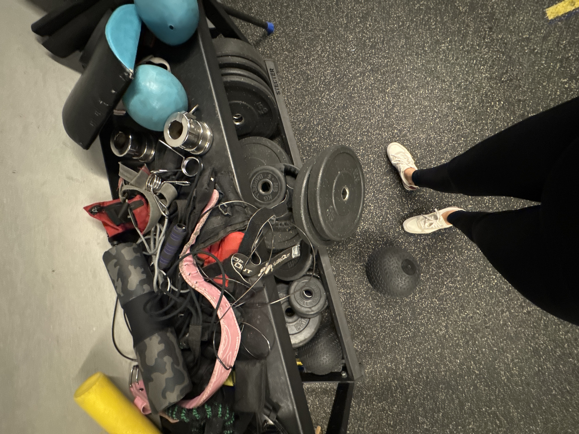 The image depicts a messy collection of gym equipment on and under a black metal weight rack. On the top shelf, a yellow foam pool noodle lies next to a rolled-up black and grey camouflage-patterned mat. Various other gym accessories, including ropes, straps, and what appear to be red gym bag components, are piled haphazardly. Several chrome-plated barbell collars are also present. To the right, two light blue exercise balls and a black piece of equipment, possibly a weightlifting belt or padding, are visible. The lower shelf of the rack is filled with numerous weight plates, some stacked and some leaning. A black slam ball rests on the grey speckled floor to the left of the rack. In the foreground, a person's legs are visible, wearing black leggings and white athletic shoes. The scene appears to be an indoor gym environment. There is no discernible indication of the city of Sochi, Russia.