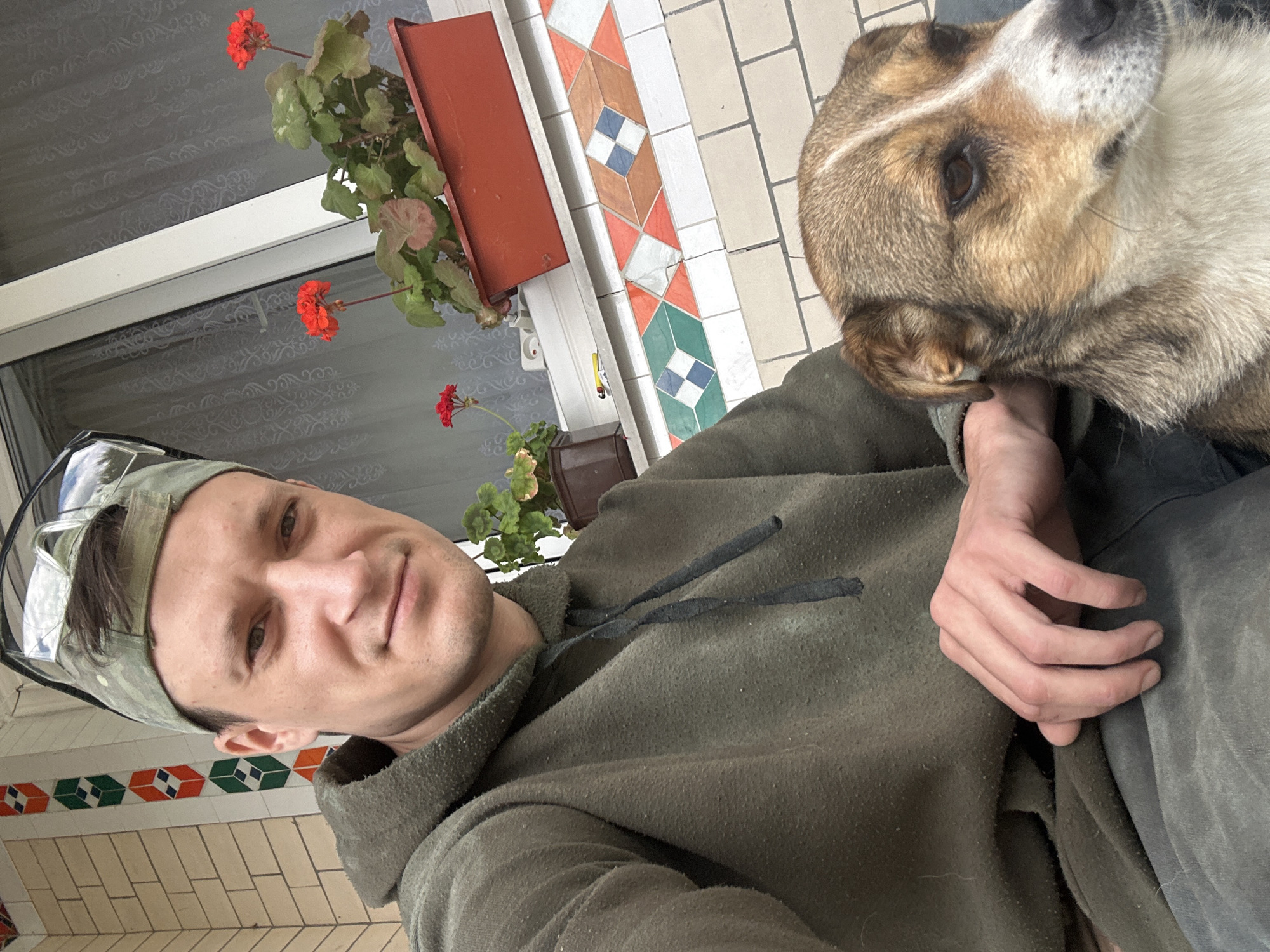 A man in a green hoodie and a camouflage baseball cap with glasses perched on top is taking a selfie with his dog on a porch. The dog, a medium-sized mixed breed with brown and white fur, is sitting beside him. In the background, there are potted red geraniums on a windowsill, a white window frame, and a wall with light-colored brick and a decorative tile pattern. The location is identified as Makiv, Ukraine.