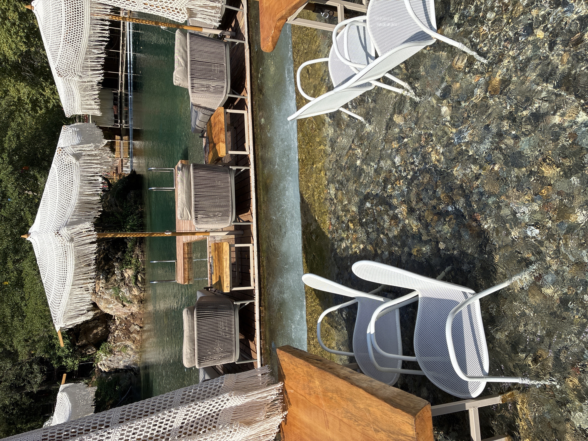 The image depicts an outdoor seating area adjacent to a clear body of water, likely a river or a pool, situated in a natural, lush environment. In the foreground, several white metal chairs with mesh seats and backs are partially submerged in shallow water with a rocky, pebbled riverbed. To the left, a thick, raw-edged wooden table rests on metal legs. In the midground, a wooden deck extends over the water, supporting several comfortable-looking lounge chairs with grey cushions. These chairs are positioned beneath large, white crocheted umbrellas with fringe detailing. Beyond the deck, the water continues, leading to rocky formations and dense green foliage characteristic of a riverside or forested area. The lighting suggests a sunny day. While the image does not explicitly show Alanya, Turkey, the presence of such a natural water feature with resort-style amenities is consistent with the tourist offerings in the region.