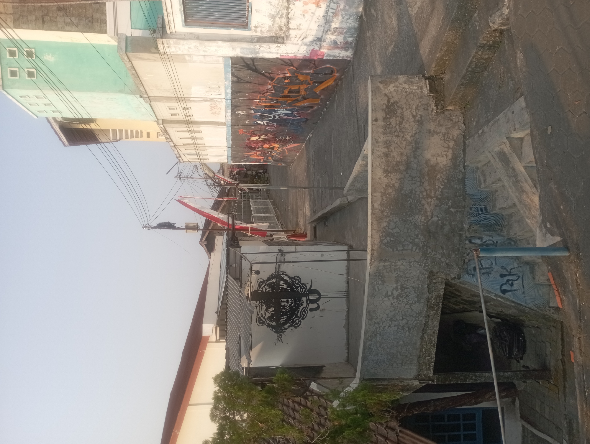 The photograph depicts a narrow alleyway in Yogyakarta, Indonesia, characterized by its urban decay and vibrant graffiti art. On the right, a wall is adorned with colorful graffiti, depicting abstract designs and stylized lettering. Adjacent to this wall, a multi-story building stands, its facade painted in alternating sections of light green and beige. Above the alleyway, electrical wires are strung across, suggesting the presence of a bustling city. On the left side of the alley, a white structure with a corrugated iron roof is visible, featuring a striking black mural of a bear's head with chains. Further left, a small building with a brown roof and a blue-framed window is partially visible, partially obscured by a tree. The ground is paved with concrete and interlocking blocks. In the foreground, concrete steps lead down, and a metal railing runs along the edge. The scene is illuminated by natural light, suggesting it is daytime.