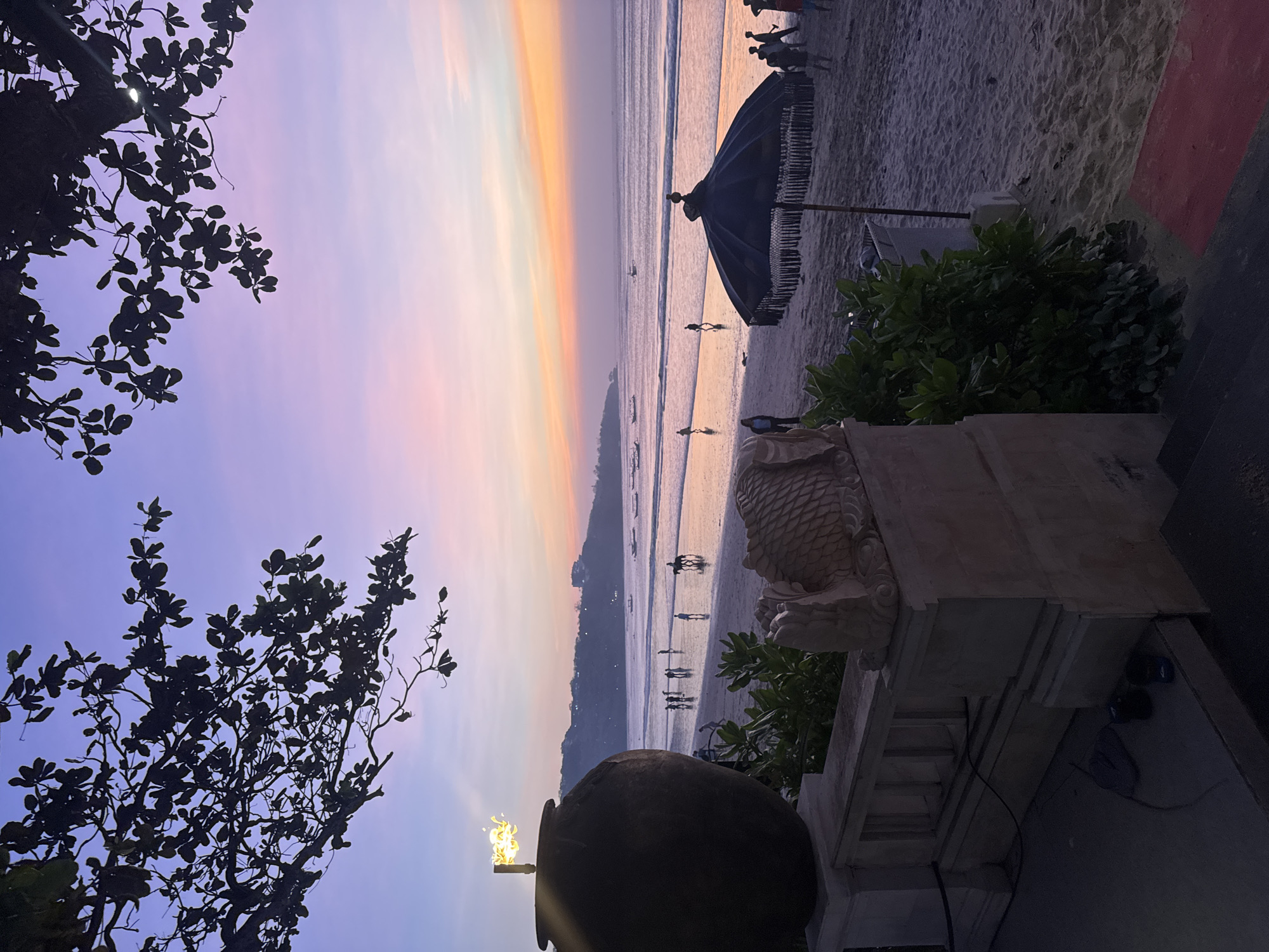 A serene beach scene unfolds at dusk in Kuta Selatan, Indonesia, with the sky painted in soft hues of purple, pink, and orange. Silhouetted branches of a tree frame the upper portion of the image, casting a dark shadow against the twilight sky. In the foreground, a large, dark urn with a flaming torch stands to the left, while a carved stone sculpture of a fish adorns a stone ledge. Lush green bushes sprout from behind the ledge, adding a touch of vibrant life to the scene. The sandy beach stretches out towards the gentle waves of the ocean, where several people are scattered, some walking along the shore, others standing near the water's edge. A figure on horseback rides along the wet sand, and a dark, fringed umbrella stands open, casting a shadow on the beach. Small boats are visible in the distance, dotting the calm sea, and a faint outline of hills lines the horizon. The overall atmosphere is peaceful and tranquil, capturing the beauty of a tropical sunset.