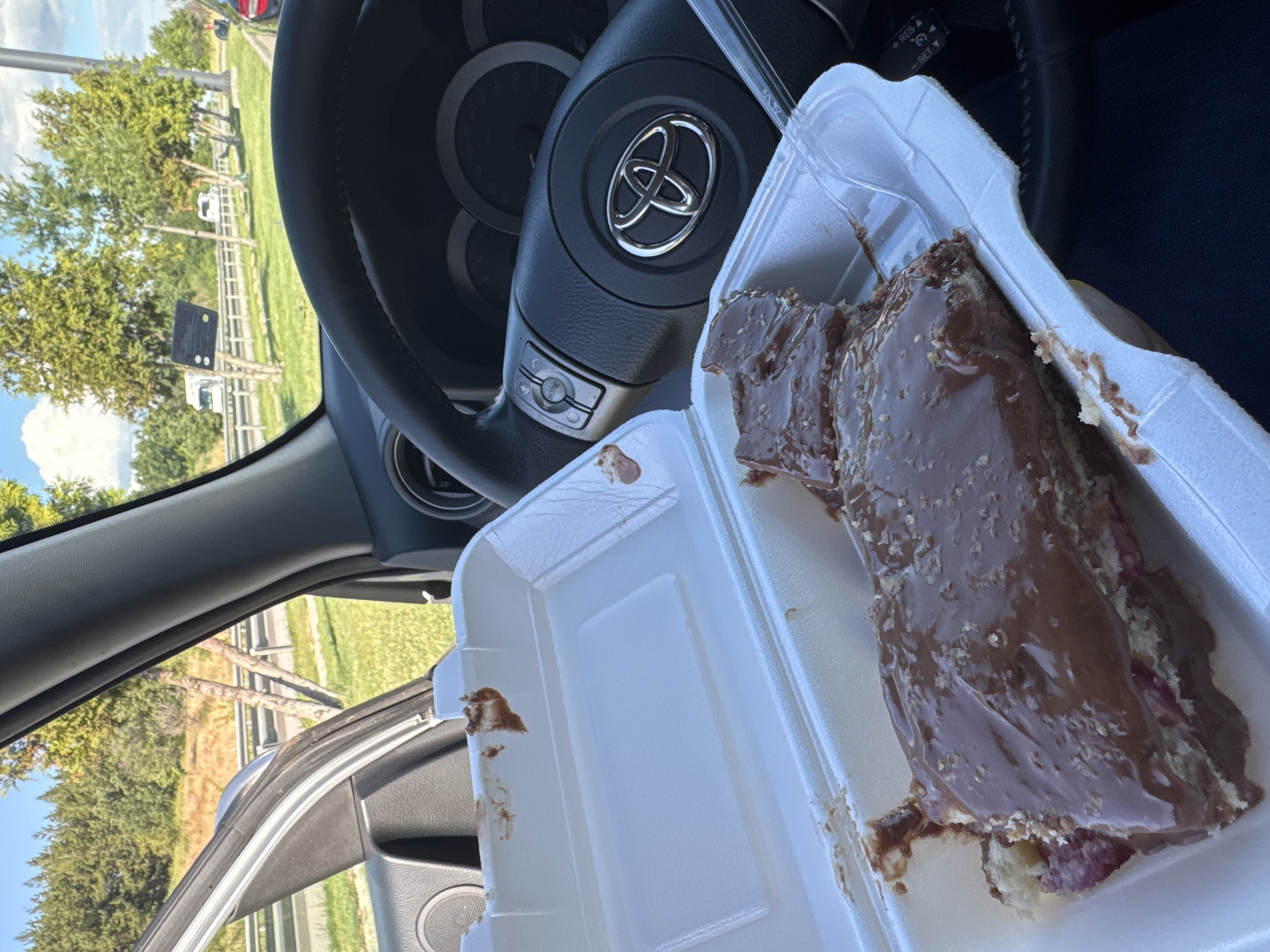 A close-up, eye-level shot shows a piece of chocolate-covered cake in a white foam takeout container, placed on the passenger seat of a car. The cake is layered and appears to have some pink filling or fruit. A clear plastic fork rests on the edge of the container. The car's dashboard and steering wheel are visible to the right, with a Toyota logo prominently displayed on the steering wheel. Outside the car window, a grassy embankment and trees line a highway, with several white trucks driving on it. The sky is blue with scattered clouds. The scene suggests the cake was recently purchased and is being enjoyed during a car journey.