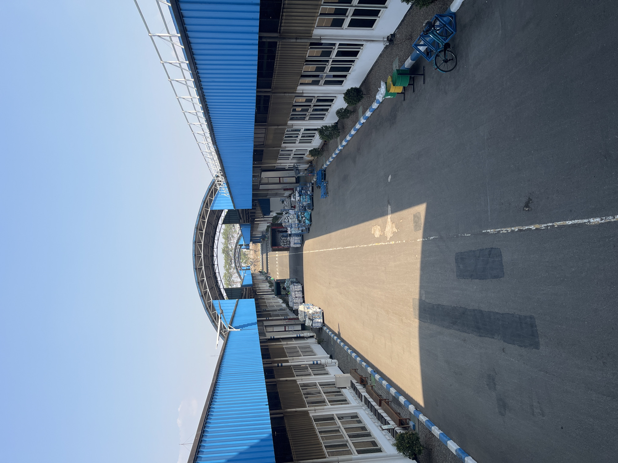 This is a high-angle, full-shot image of an industrial complex with a clear blue sky. The complex features long, rectangular buildings with blue corrugated metal roofs and tan-colored corrugated metal siding. White-framed windows are visible along the buildings. A paved road runs between the two rows of buildings, with a distinct line of blue and white striped curbing along the edges of the road. Stacked pallets of goods are visible on the sides of the road, some wrapped in plastic. In the distance, more buildings with similar blue roofs are visible, connected by arched structures. On the right side of the road, there are green and yellow cylindrical trash bins and a blue bicycle rack with a bicycle. The ground of the road has some darker patched areas and white lines marking lanes. There are no people visible in the image. The environment appears to be outdoors in a sunny climate. The location is identified as the city of Pelang, Indonesia.