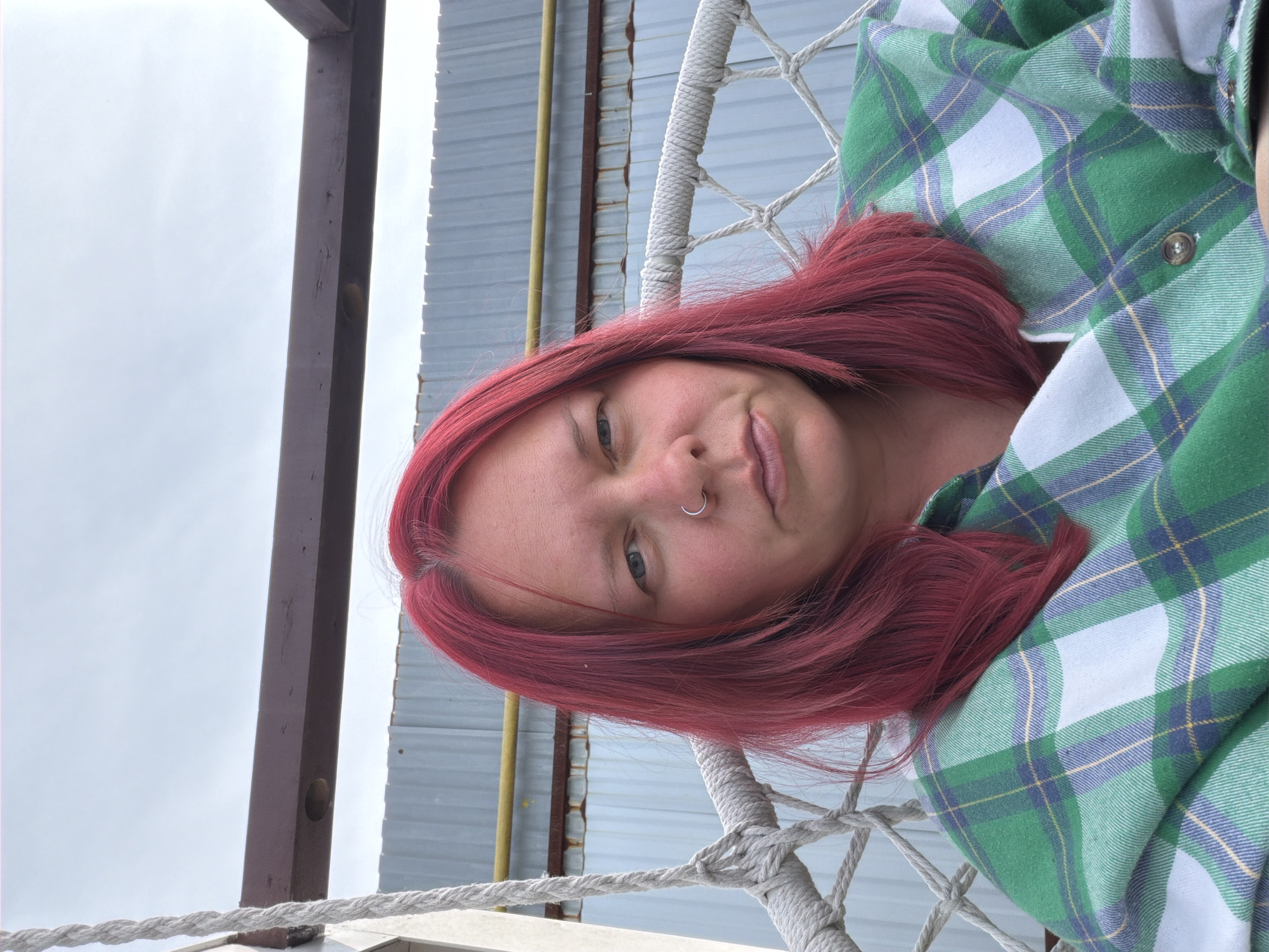 A young woman with red hair and a nose ring is lying in a rope hammock. She is wearing a green and white plaid shirt. The hammock is suspended from a wooden structure. Behind the structure is a corrugated metal wall. The sky is overcast. This image may have been taken in an outdoor setting, possibly in Chaikovsky, Russia.