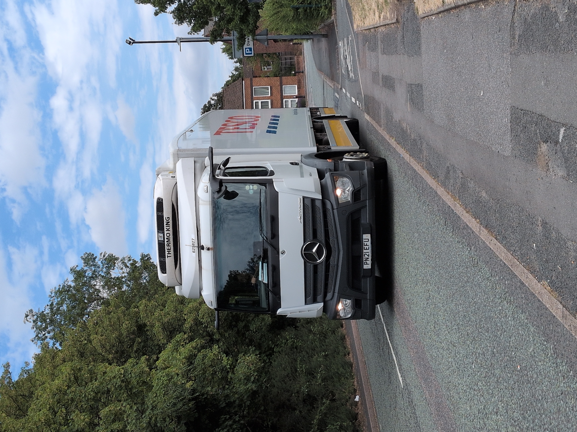 A white Tesco truck with the license plate PN21 EFU is driving on a road in Dawley, United Kingdom. The truck is a Mercedes-Benz Actros and has a Thermo King refrigeration unit on top. The truck is in motion, as indicated by the blur of the wheels and the motion lines on the road. The road is asphalt and appears to be in good condition. There are trees on the left side of the road and buildings on the right side. The sky is blue with some clouds. There are no people visible in the image.