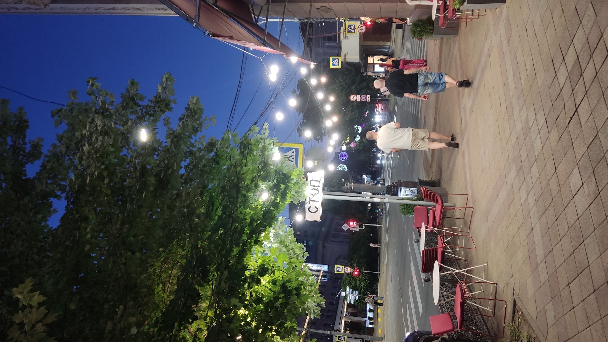 This image depicts an urban street scene in Krasnodar, Russia, during the evening. A tree with lush green foliage overhangs the sidewalk on the left. String lights are suspended across the street, casting a warm glow. A stop sign, written in Russian ("СТОП"), is prominently displayed on a pole near the sidewalk.
On the sidewalk, two men are walking away from the camera. The man on the left wears a white t-shirt and beige shorts, while the man on the right wears a black t-shirt and denim shorts. Further down the sidewalk, a woman in a pink top and patterned pants is crossing the street. Several small tables with red chairs are set up on the sidewalk, suggesting an outdoor seating area for a cafe or restaurant.
The street itself is paved, with a pedestrian crossing visible in the foreground. Traffic lights are illuminated, and cars are visible on the street, their headlights on. The buildings lining the street appear to be multi-story structures, some with illuminated windows. The sky is a deep blue, indicating dusk or early night. The overall impression is a bustling city environment transitioning into the evening.