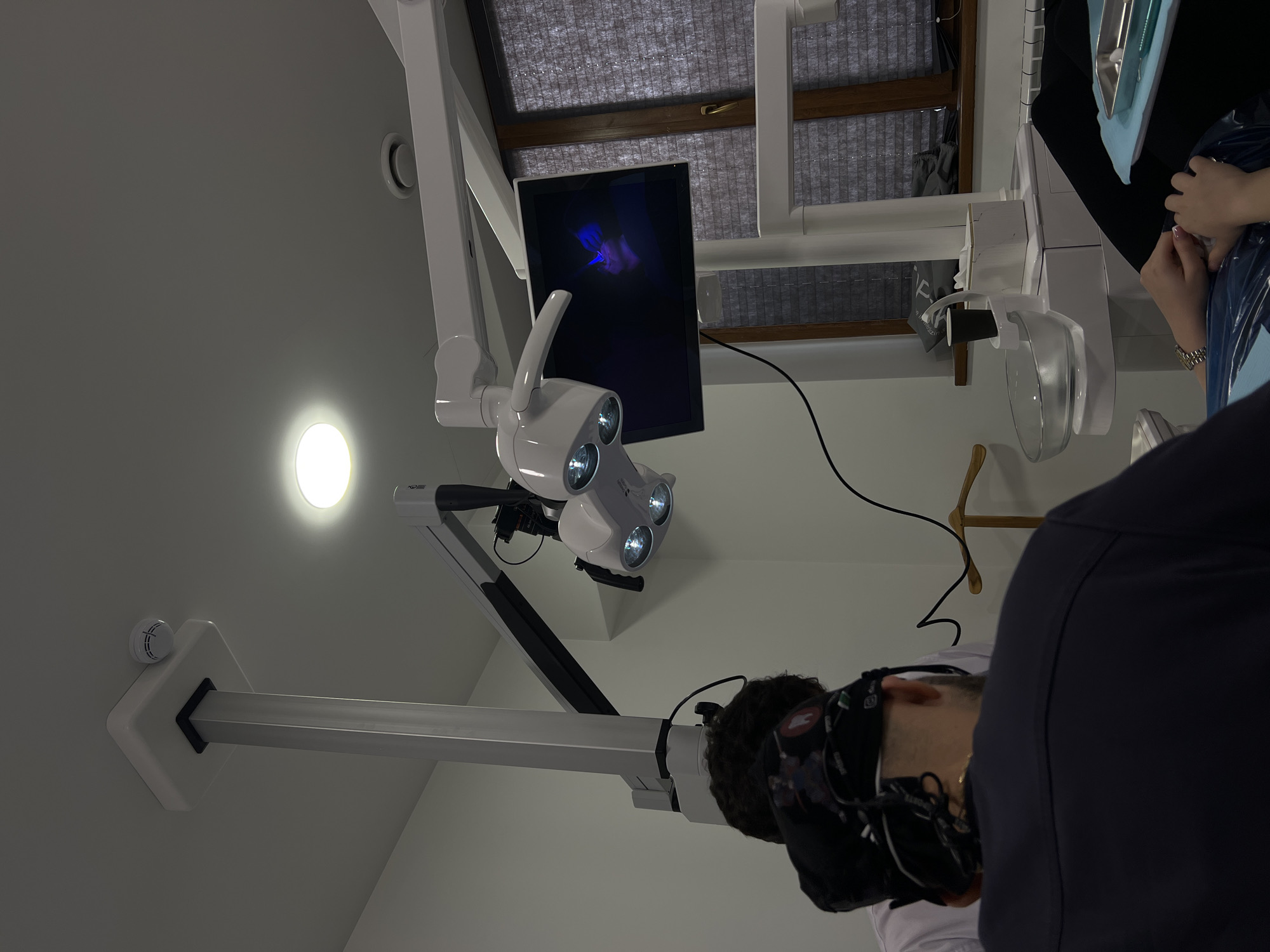 The image depicts a dental examination room. A dental examination light is suspended from the ceiling, directly above a patient lying in a dental chair. A screen is visible behind the light, displaying an image. An individual, presumably a dentist or assistant, is partially visible in the bottom right, wearing dark clothing. The environment is brightly lit, clean, and sterile. The location is in a modern dental office, possibly in Yerevan, Armenia.