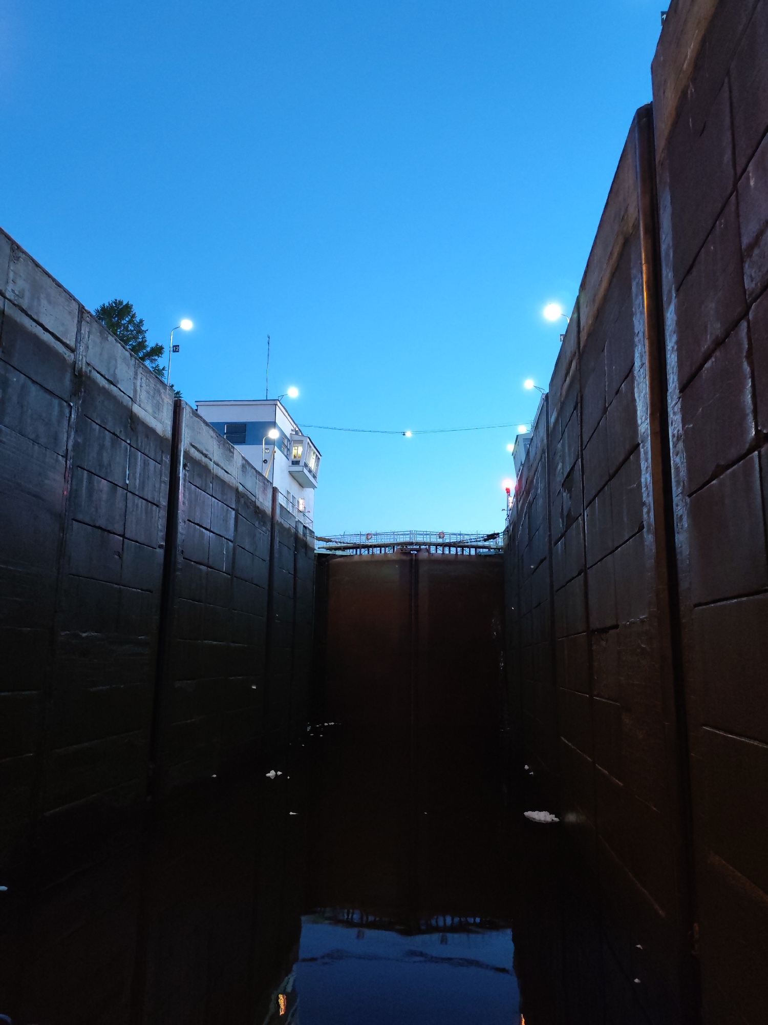 The image captures a narrow view into a large, dark lock chamber during twilight or early dawn. Two imposing, tall concrete walls, appearing wet and textured, rise steeply on either side, creating a confined passageway. The water at the bottom of the chamber is dark and reflective, mirroring the deep blue sky above and the bright artificial lights.
At the far end of the lock, large, dark gates are visible, suggesting the chamber is currently closed. Overhead, the sky transitions from a lighter blue near the horizon to a deeper, darker blue at the zenith. Several bright, white streetlights or floodlights illuminate the top edges of the walls and the surrounding area. Smaller lights are also strung across the width of the lock further down the chamber.
On the left side, above the concrete wall, a multi-story white building with windows is visible, likely a control tower or lock house, also lit up. Nearby, a small white sign on a pole reads "12". A few trees are silhouetted against the sky behind the left wall. The right wall also shows industrial structures and railings along its top edge. The overall atmosphere is quiet and industrial, with the artificial lights contrasting against the natural twilight sky and the dark, imposing structures of the lock. No people are visible, but the scene implies the functionality of a waterway for navigation.