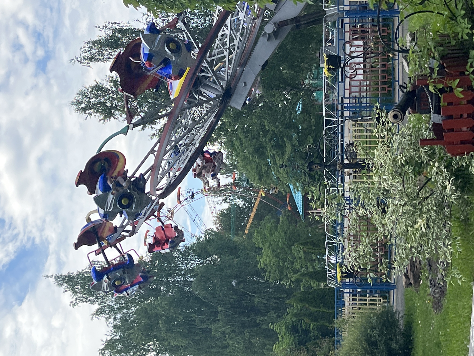 The image depicts a section of an amusement park in Chaikovsky, Russia, under a cloudy sky. In the foreground, a decorative red fence encloses a small, antique-looking cannon. Beyond this, a vibrant green lawn is dotted with plants, and a large, leafy bush with variegated green and white leaves partially obscures the view.
Dominating the mid-ground and background are several amusement rides. A large, angular ride with metallic arms and brightly colored seats, featuring red and blue elements, is prominently positioned on the right side of the frame, angled upwards. Passengers are visible in some of the seats. To the left and further back, a Ferris wheel with red gondolas can be seen. Smaller amusement rides with various structures and colored elements are scattered throughout the park.
The park is surrounded by a dense growth of tall, mature trees, primarily deciduous with full green canopies, some with lighter green leaves suggesting birch trees. Behind the rides, a few smaller, conical evergreen trees are visible. A multi-colored fence, with horizontal bars in blue, yellow, and red, runs across the mid-ground, separating different areas of the park. Parts of buildings with teal-colored roofs are visible between the rides and trees. The overall impression is of a lively, well-established amusement park on a day with mixed weather conditions.