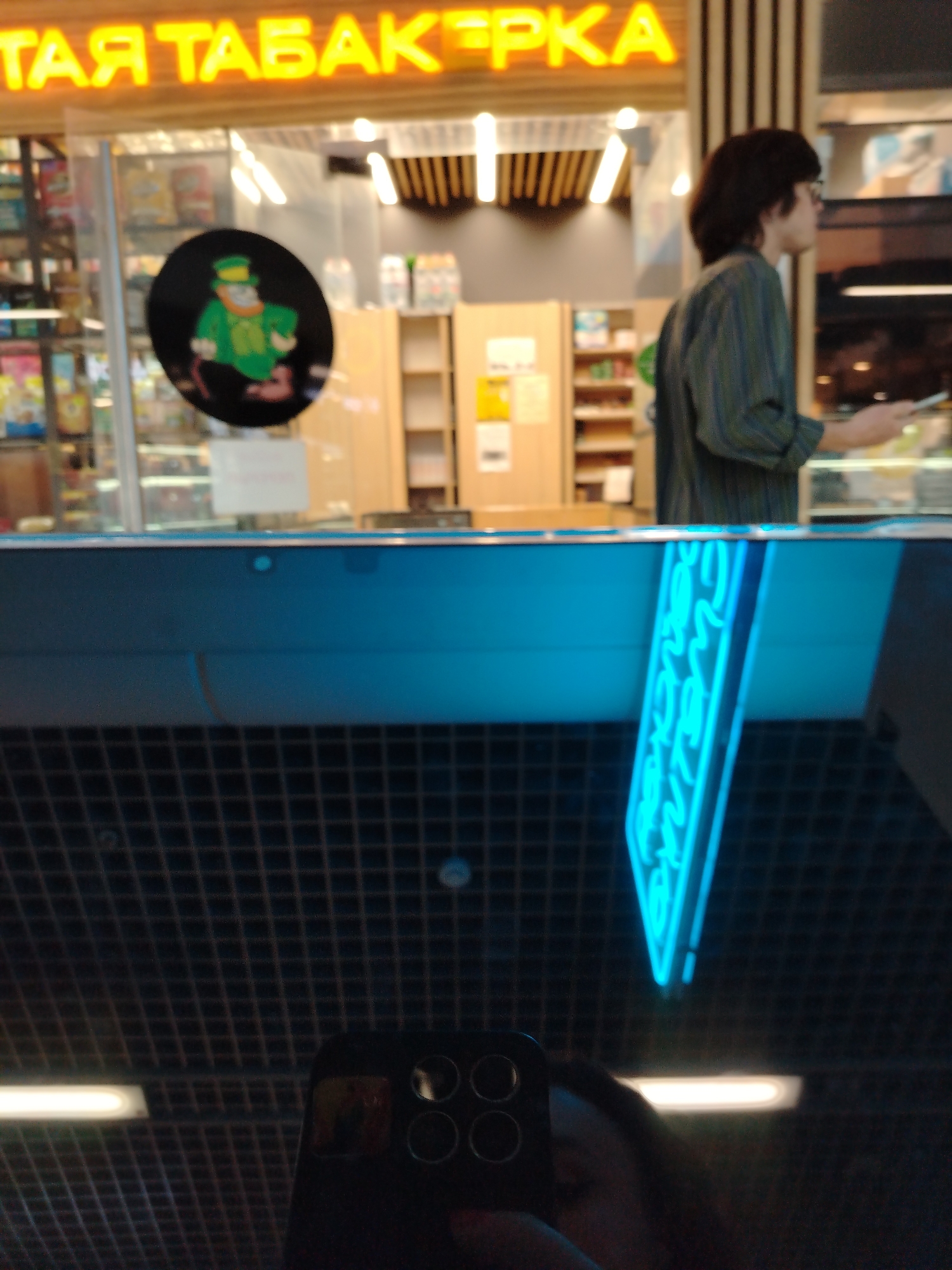 The image shows a reflection of a store interior on a glass surface. In the background, a person with dark hair and glasses wearing a striped shirt is visible. Above them, a neon sign with Russian text is illuminated. A round sticker with a cartoon leprechaun is affixed to the glass. In the foreground, a smartphone is being held, capturing the reflection. The scene suggests a commercial environment, possibly a shop or kiosk, within a public space like a mall. The neon sign and the type of store indicated by the leprechaun sticker point towards a tobacco shop. Based on the Russian text on the sign, it is likely located in Russia, potentially in Izhevsk given the provided context. The overall impression is a casual snapshot taken by a visitor or customer.