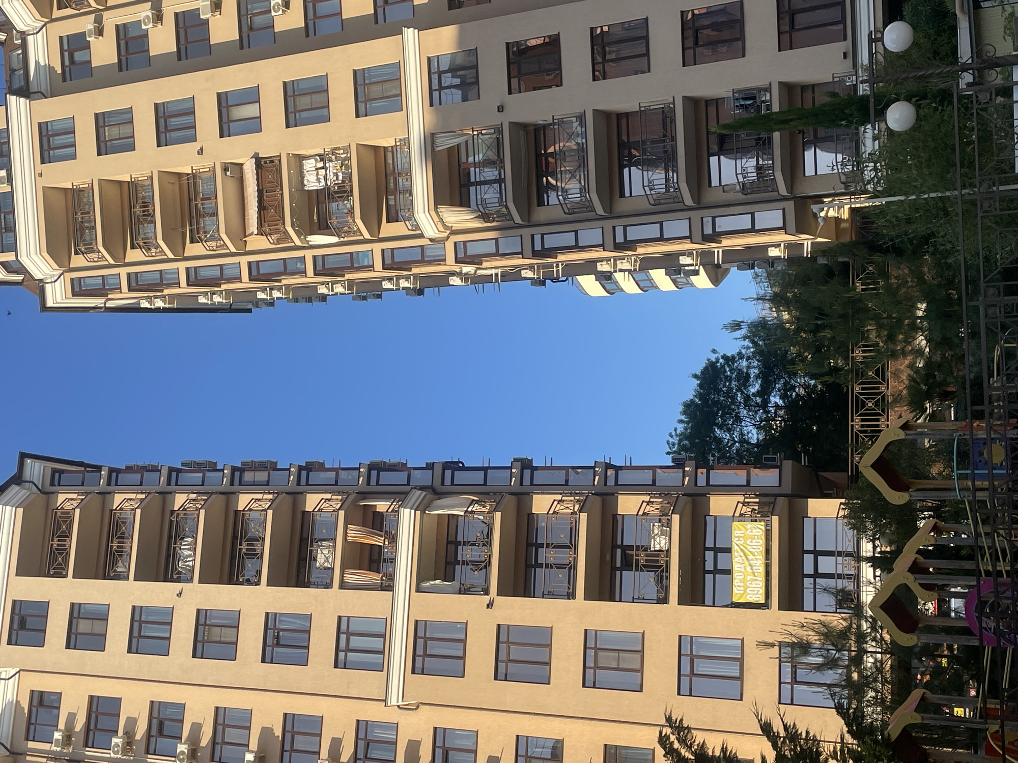 This image captures a view between two modern, multi-story residential buildings in the city of Sochi, Russia, under a clear blue sky. Both buildings feature a light beige or tan facade with numerous brown-framed windows and curved balconies adorned with decorative metal railings.
The building on the left shows several balconies equipped with external air conditioning units. On one balcony, white fabric or clothes are hanging to dry, while another shows beige curtains. Towards the lower floors, a large yellow banner is prominently displayed on a balcony, reading "ПРОДАЕТСЯ" (FOR SALE) with a phone number "8967-641-06-67" below it. Below this building, a children's playground with colorful structures (including red, yellow, and purple elements) is visible, surrounded by lush green trees and shrubs.
The building on the right mirrors the architectural style, also featuring multiple balconies with similar railings and brown-framed windows. Some balconies have visible items such as curtains, potted plants, or small pieces of furniture. Air conditioning units are also mounted on its exterior. At ground level, dense green foliage, tall slender trees, and spherical white outdoor lights are discernible, partially obscured by a dark metal fence in the foreground.
The overall scene is bathed in sunlight, highlighting the architectural details and the vibrant greenery, consistent with a warm, temperate climate. No people are visible in the image.