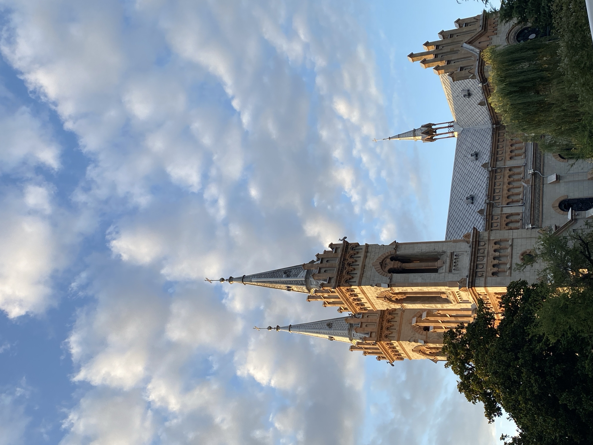 The image depicts the Holy Trinity Cathedral of Batumi, Georgia, on a sunny day with scattered clouds. The cathedral is a prominent religious building with two tall spires topped with crosses. The facade is made of light-colored stone with intricate architectural details, including arched windows and decorative elements. Green trees and foliage surround the base of the cathedral, partially obscuring the lower levels. The sky is a bright blue, with fluffy white clouds that are catching the sunlight, suggesting it is either morning or late afternoon. The overall impression is one of a majestic religious structure set against a beautiful natural backdrop.