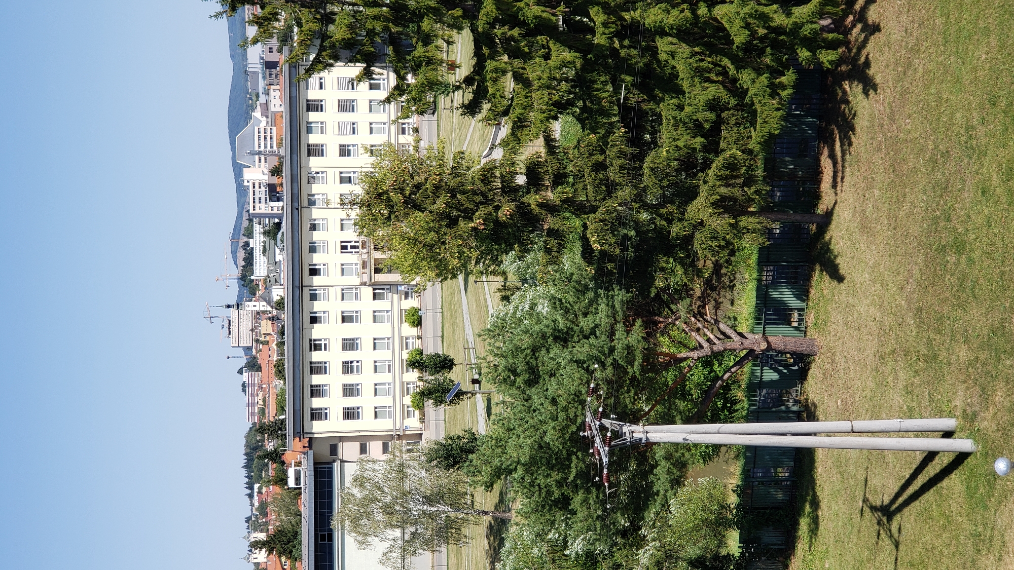 The image captures a view of a modern building complex in Prešov, Slovakia, under a clear blue sky. The foreground features a well-maintained grassy area with shadows cast by trees and a pole with electrical insulators. A green metal fence runs along the middle ground, separating the lawn from a wooded area with mature coniferous trees. In the background, a long, light-colored building with numerous windows stands prominently, suggesting it is an institutional or office building. Beyond this, a cityscape is visible with a mix of residential and commercial structures, along with construction cranes, indicating ongoing development. The overall scene is one of urban landscape bathed in daylight, devoid of any human presence.