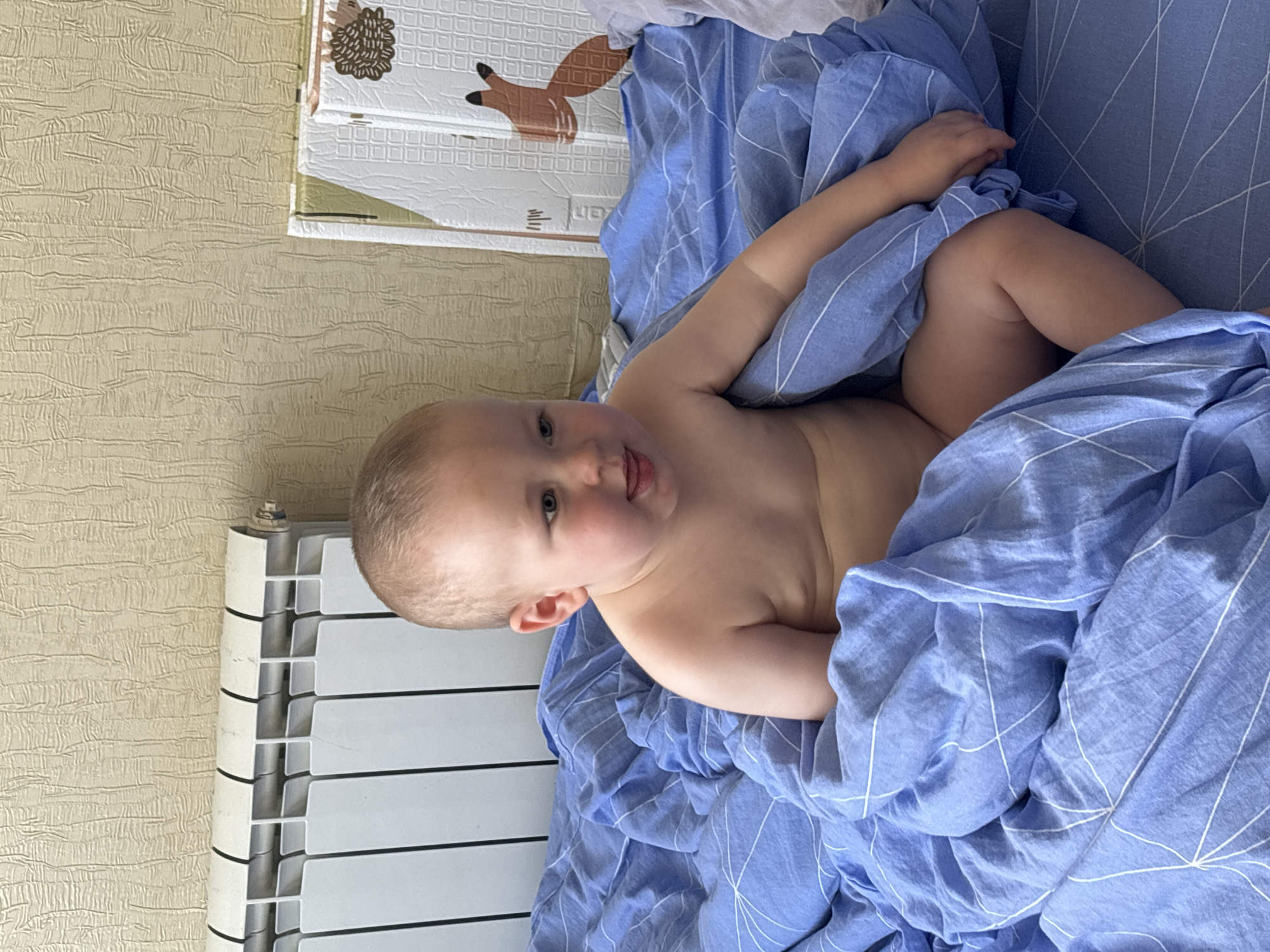 A baby is partially covered by a blue comforter with white lines on a bed. The baby is looking at the camera and sticking out their tongue. The baby's head and shoulders are visible. The setting appears to be a bedroom, with a radiator to the left of the frame and a textured wall behind it. A play mat with animal illustrations is visible in the background. The image is taken indoors.