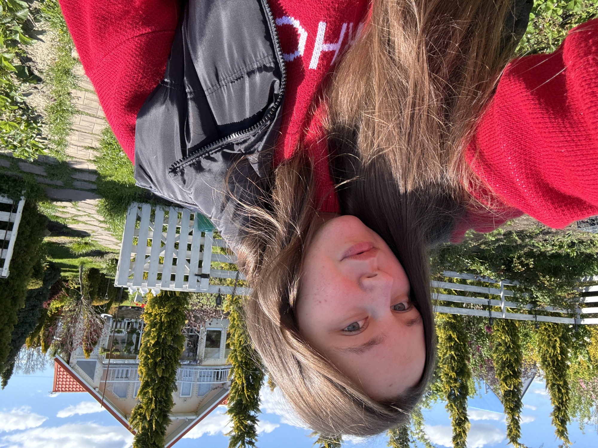 An outdoor, upward-facing selfie captures a young woman in bright daylight, likely in Pervorechenskoye, Russia. Her face, positioned towards the right side of the frame, shows light-colored eyes and a neutral expression. Long, light brown hair cascades around her face and shoulder. She is dressed in a red sweater or hoodie with white capital letters "OH" visible on the fabric, layered with a black puffer vest featuring a visible zipper.
The background depicts a vibrant garden or yard. A white picket fence stretches across the middle ground, partially bordering a paved walkway or path that meanders through green grass and various plants. Further back, a multi-story house with a distinctive red-tiled roof, white siding, and possibly a balcony stands amidst several tall, slender evergreen trees and other deciduous foliage. The sky above is clear and blue with a few scattered white clouds, indicating a sunny day. The scene evokes a tranquil, residential atmosphere.