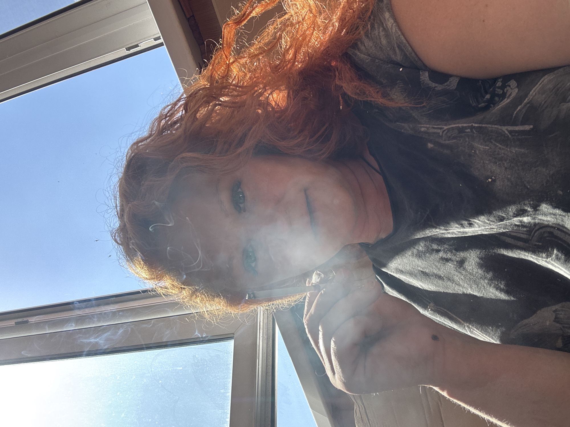 A woman with red hair is holding a lit cigarette near a window, with smoke rising. The view outside the window shows a clear blue sky. The woman is wearing a grey t-shirt with a pattern. She has fair skin and blue eyes. The location is Chaikovsky, Russia.