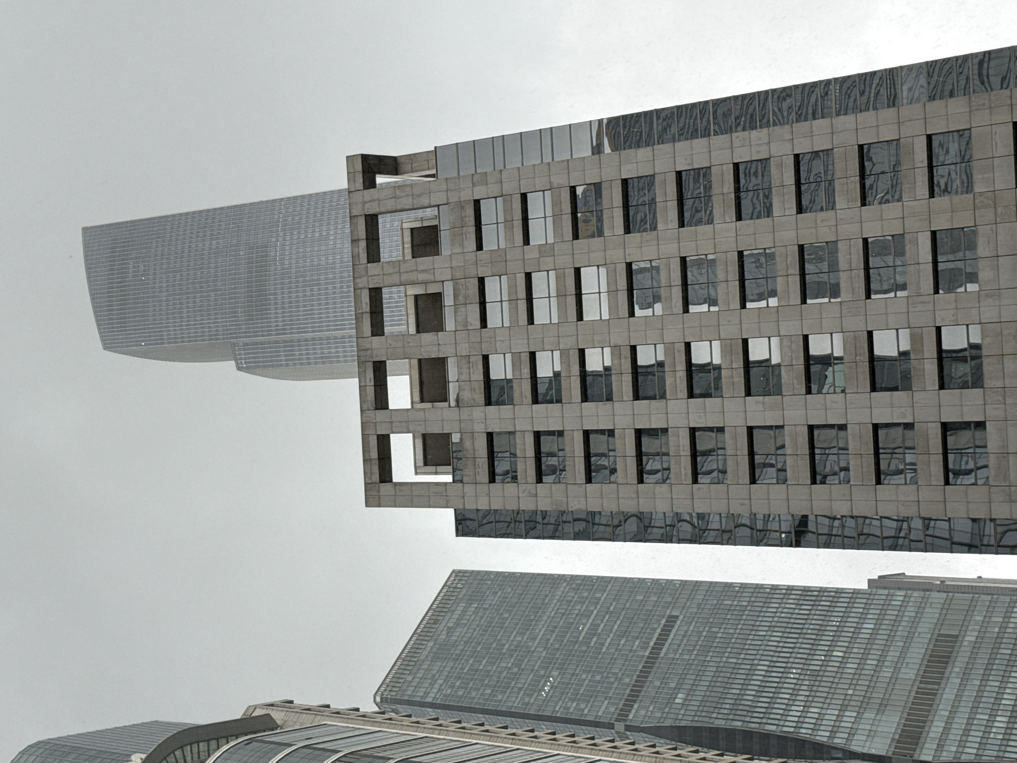 This is a low-angle, full shot of a cluster of modern skyscrapers under a grey, overcast sky, likely in Tianhe District, China. In the foreground and to the right, a tall building with a gray, textured facade is visible, featuring rows of dark, rectangular windows. To the left and slightly behind this building, a much taller, slender skyscraper with a predominantly glass facade dominates the frame. Its surface is a mosaic of vertical and horizontal lines, reflecting the overcast sky. Further in the background, another immense skyscraper, lighter in color and with a curved silhouette, rises into the sky. Its surface appears to be composed of numerous vertical strips. The overall impression is of a dense urban environment with towering architectural structures. There are no people visible in the frame.