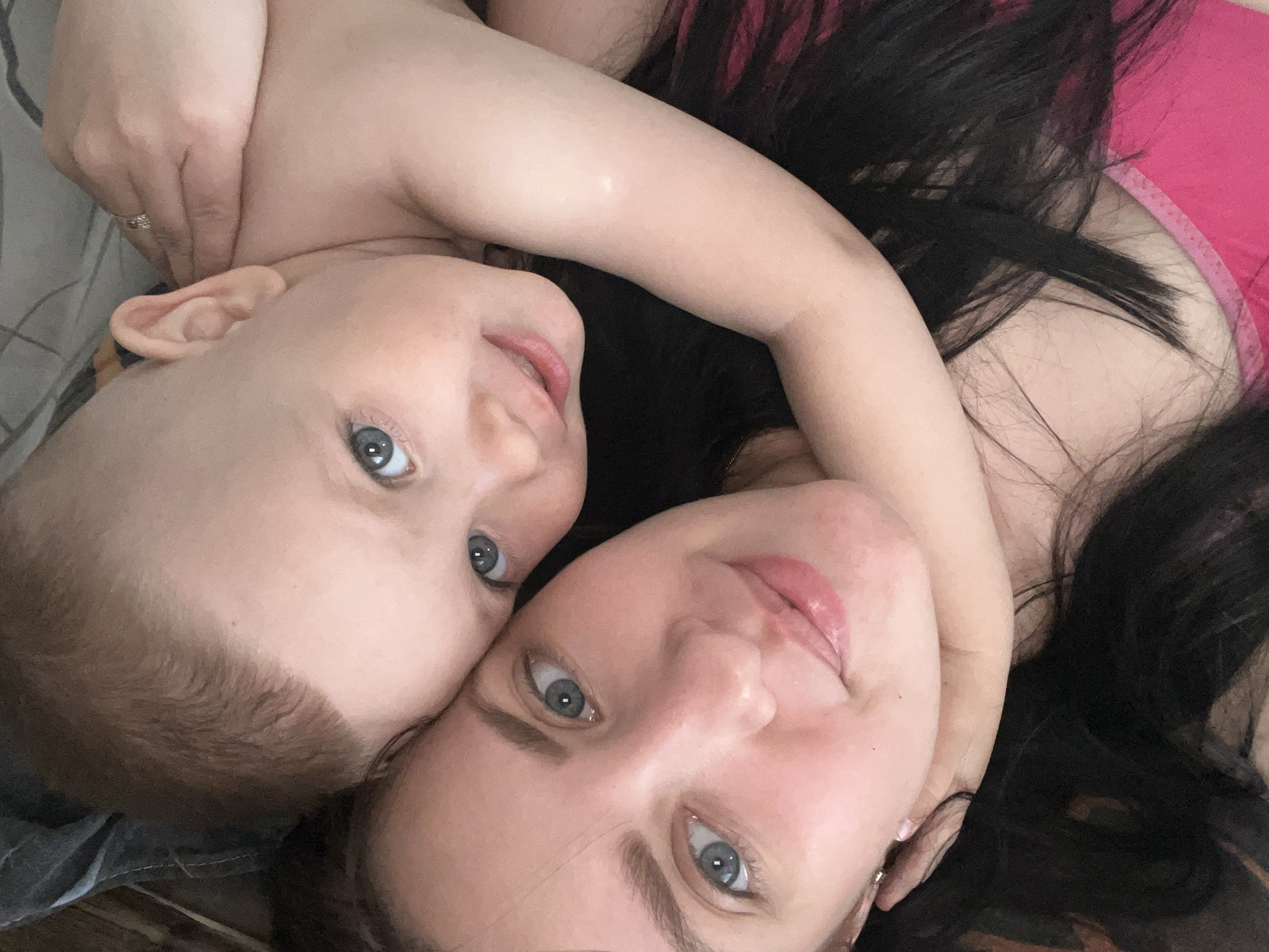 A mother and child lie closely together, facing the camera. The child, with fair skin, short brown hair, and blue eyes, has an arm around the mother. The mother has long dark hair, fair skin, and blue eyes. They appear to be indoors. The location is Марковский, Russia.
