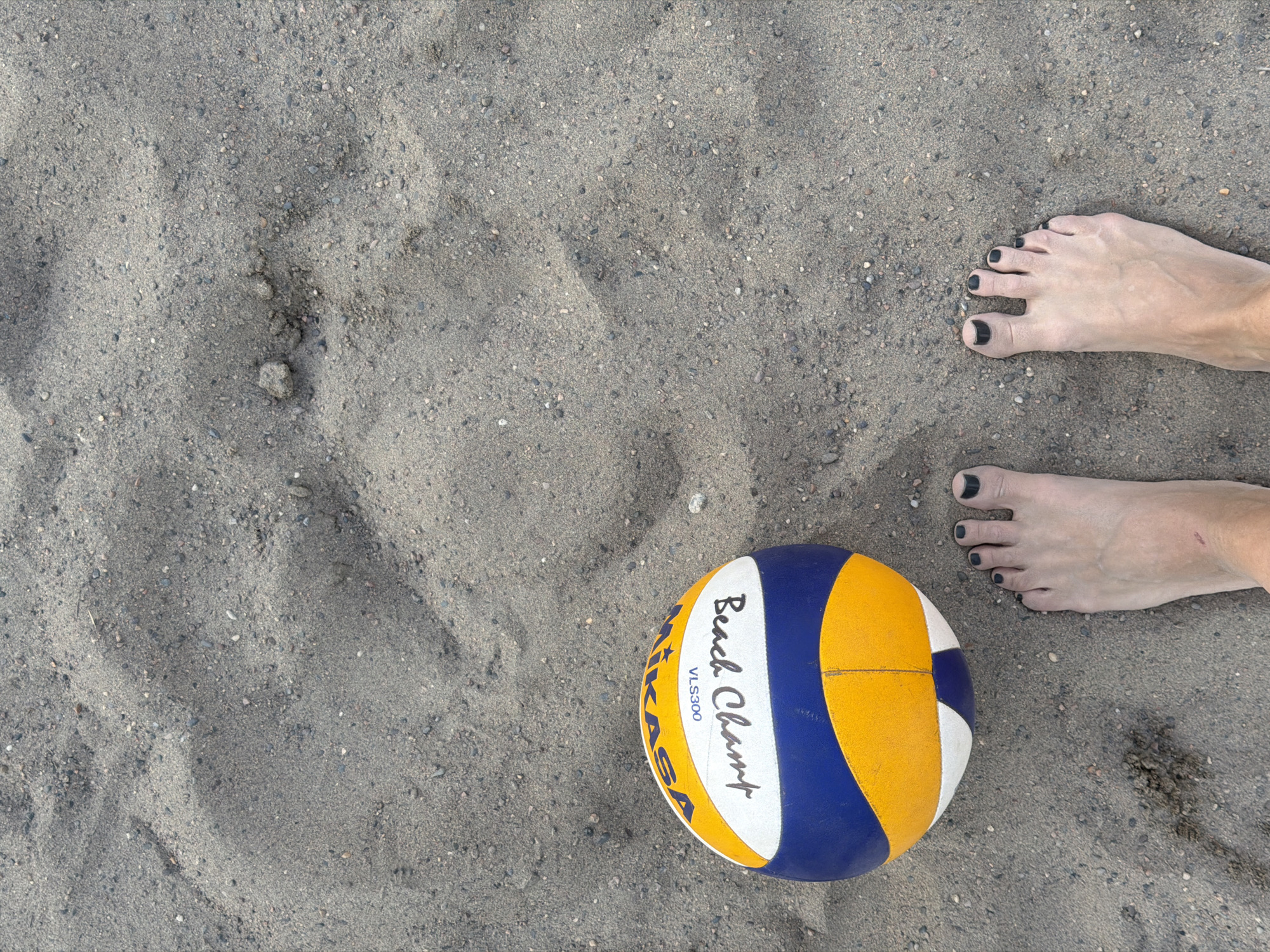 This image presents a top-down view of a sandy surface, featuring human feet and a volleyball. The predominant subject is the sand, a light gray-brown texture mixed with numerous small pebbles and showing subtle ripples and indentations.
In the upper right and lower right portions of the frame, a pair of bare feet are visible, likely belonging to a woman, given the dark painted toenails. One foot is positioned in the upper right, with toes pointing left, while the other is in the lower right, with toes pointing upwards and slightly left.
A blue, yellow, and white volleyball, specifically a Mikasa "Beach Champ" VLS300 model, is centrally placed in the lower right quadrant of the image. Its design features distinct colored panels.
The scene strongly implies a beach or a sandy volleyball court, suggesting an activity related to beach volleyball or a moment of rest from it. The lighting appears even and diffused, characteristic of an overcast day or a time of day with no harsh direct sunlight. Given the location context of Krasnoyarsk, Russia, the setting is likely a river beach along the Yenisei River or an artificial sandy recreation area. The overall impression is one of leisure and outdoor activity.