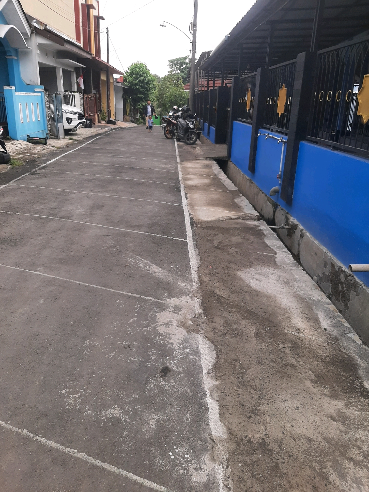 The image depicts a paved outdoor area, likely a parking lot or street, in Sumampir, Indonesia. The ground is asphalt with visible white painted parking lines. To the left, a blue and white building with an arched entrance and a white car parked in front of it is partially visible. A white fence and a gate are also present. Further down the street, two motorcycles are parked, and a man and a child are walking away from the camera. To the right, a black metal fence with decorative yellow stars and circular motifs separates the paved area from a building with a blue wall. A concrete drainage channel runs alongside the blue wall, containing pipes and a faucet. The environment appears to be urban, with buildings and vegetation visible in the background under an overcast sky.