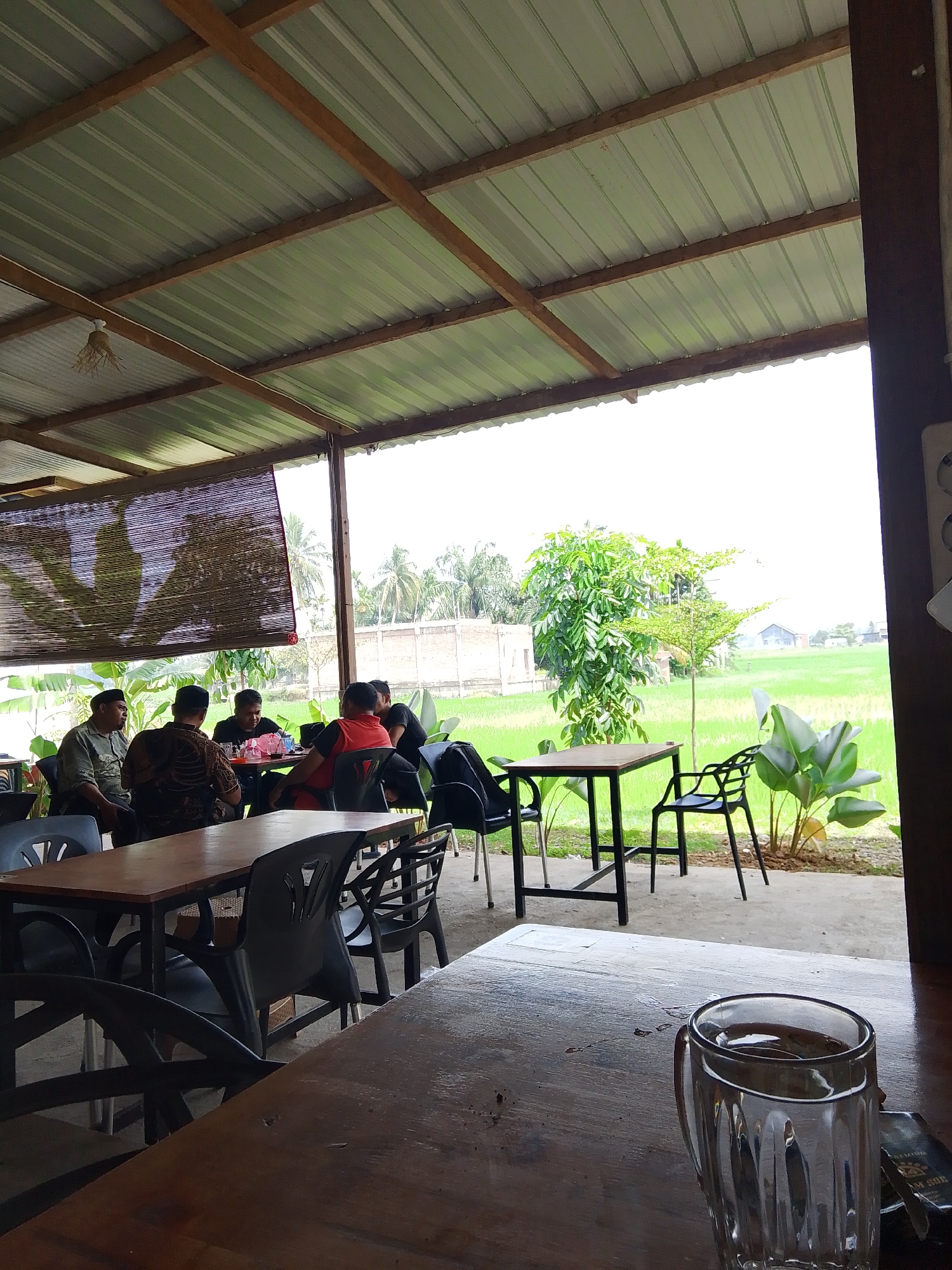 The image depicts an outdoor seating area of a cafe, likely in Meunasahmee, Indonesia, overlooking a vibrant green rice field. Several people are seated at tables, some engaged in conversation, others appearing to be eating or drinking. The architecture suggests a simple, open-air structure with a corrugated metal roof supported by wooden beams. In the foreground, a wooden table holds a glass of a dark liquid, possibly coffee or tea, and an open pack of cigarettes. The background reveals lush vegetation, including palm trees and banana plants, against a backdrop of a distant building and the expansive rice paddy. The overall atmosphere appears relaxed and casual, with natural light illuminating the scene.