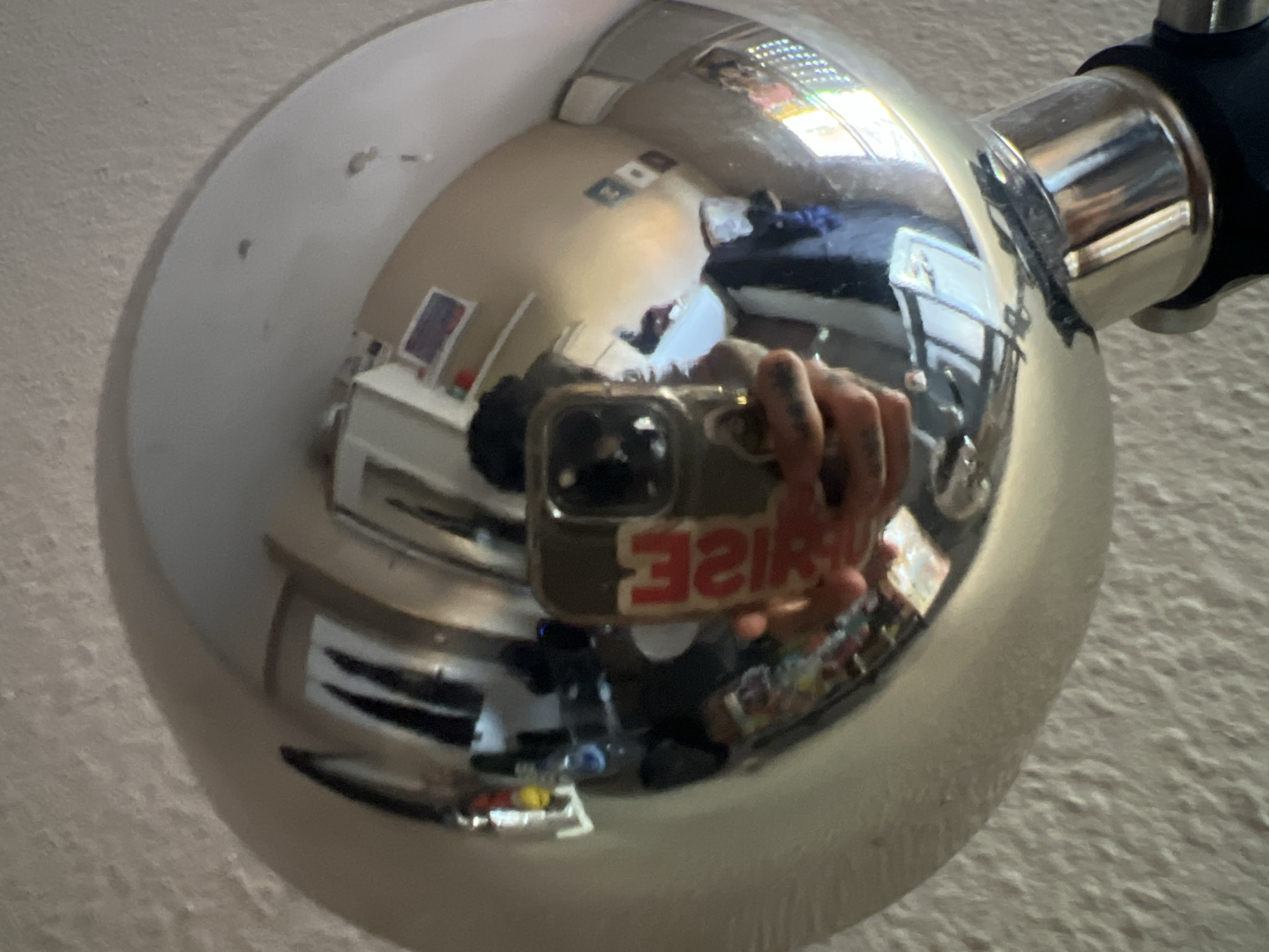 The image is a reflection in a chrome sphere, seemingly part of a handlebar-mounted mirror on a vehicle, possibly a bicycle or motorcycle. The reflection shows a bedroom interior with a bed, dresser, and window. A person is visible holding a phone with a reversed sticker visible on the case. The setting is likely indoors, based on the furniture and lighting. Location data is unavailable; therefore, a determination regarding Madrid, Spain is not possible.