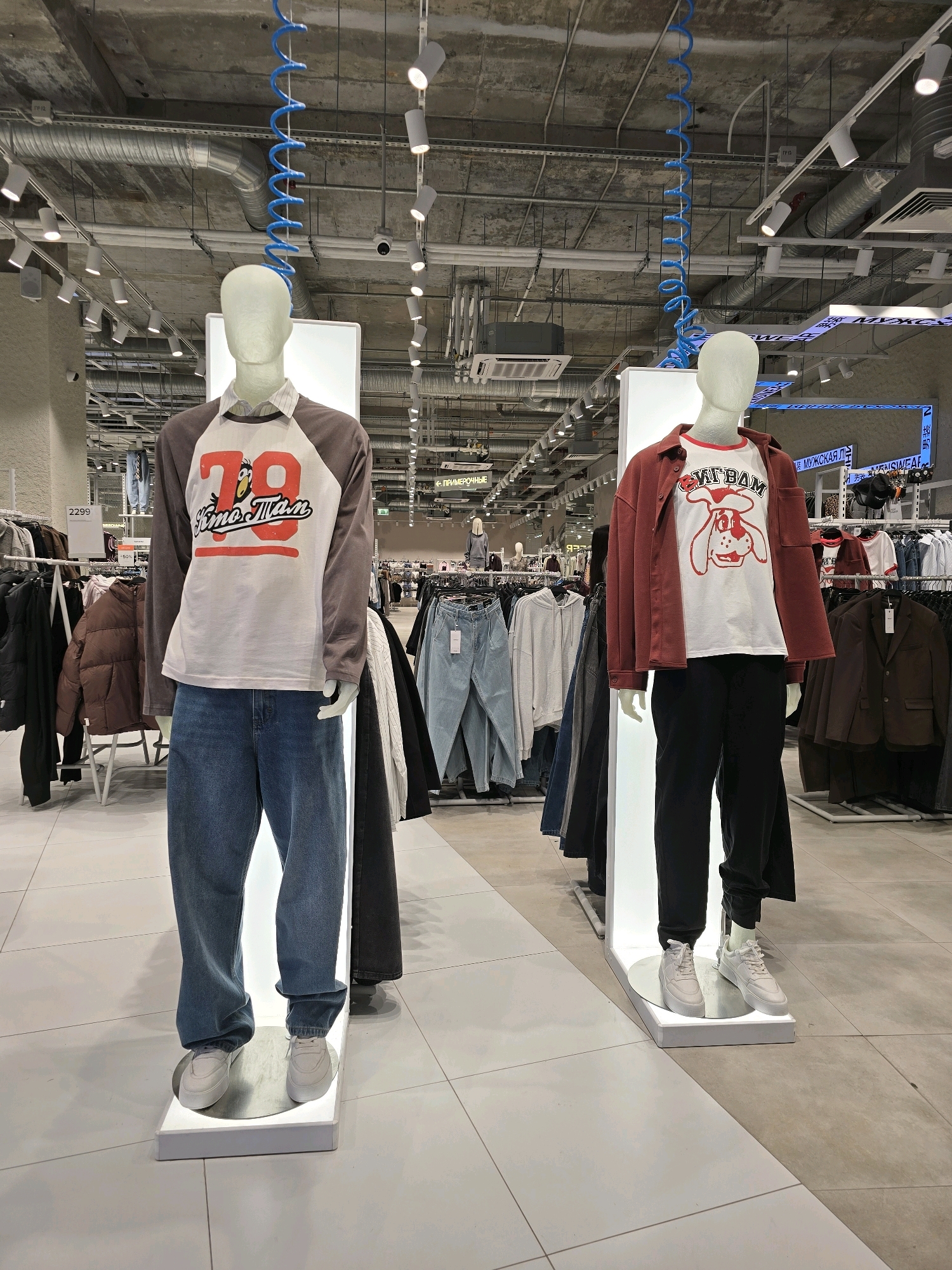 The image shows two mannequins in a retail store. The mannequin on the left is wearing a long-sleeved shirt with a gray raglan sleeve and a white front featuring a graphic of the number "78" and cartoonish text, paired with loose-fitting blue jeans and white sneakers. The mannequin on the right is dressed in a white t-shirt with a graphic of a cartoon dog's face, a rust-colored shirt jacket, black pants, and white sneakers. Both mannequins are displayed on illuminated white pedestals. The background reveals racks of clothing, store lighting, ventilation systems, and signage in Russian, including "МУЖСКАЯ" (men's). The presence of Cyrillic signage and typical retail store architecture suggests the location is likely in Russia. The store appears to be a clothing retailer.