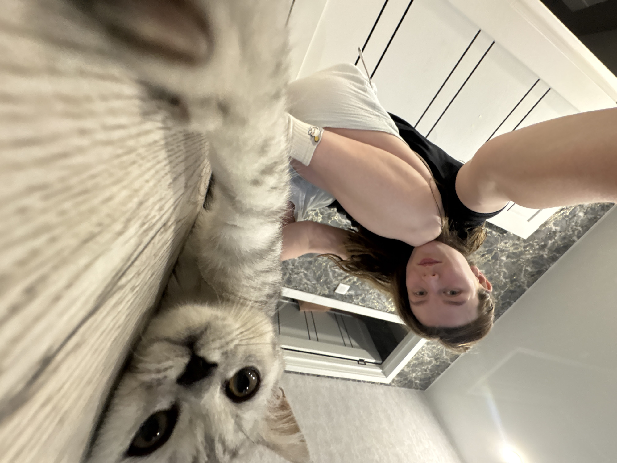 A selfie shot in a bathroom, likely in a residence, shows a young woman holding the camera at an angle, with a gray and white cat appearing prominently in the foreground. The woman has light skin and shoulder-length brown hair, and she is wearing a black top and light-colored shorts and socks. The cat is close to the camera, its large eyes in focus. The bathroom has white walls with vertical black lines and marble countertops. A window is visible in the background. The location is likely Izhevsk, Russia.
