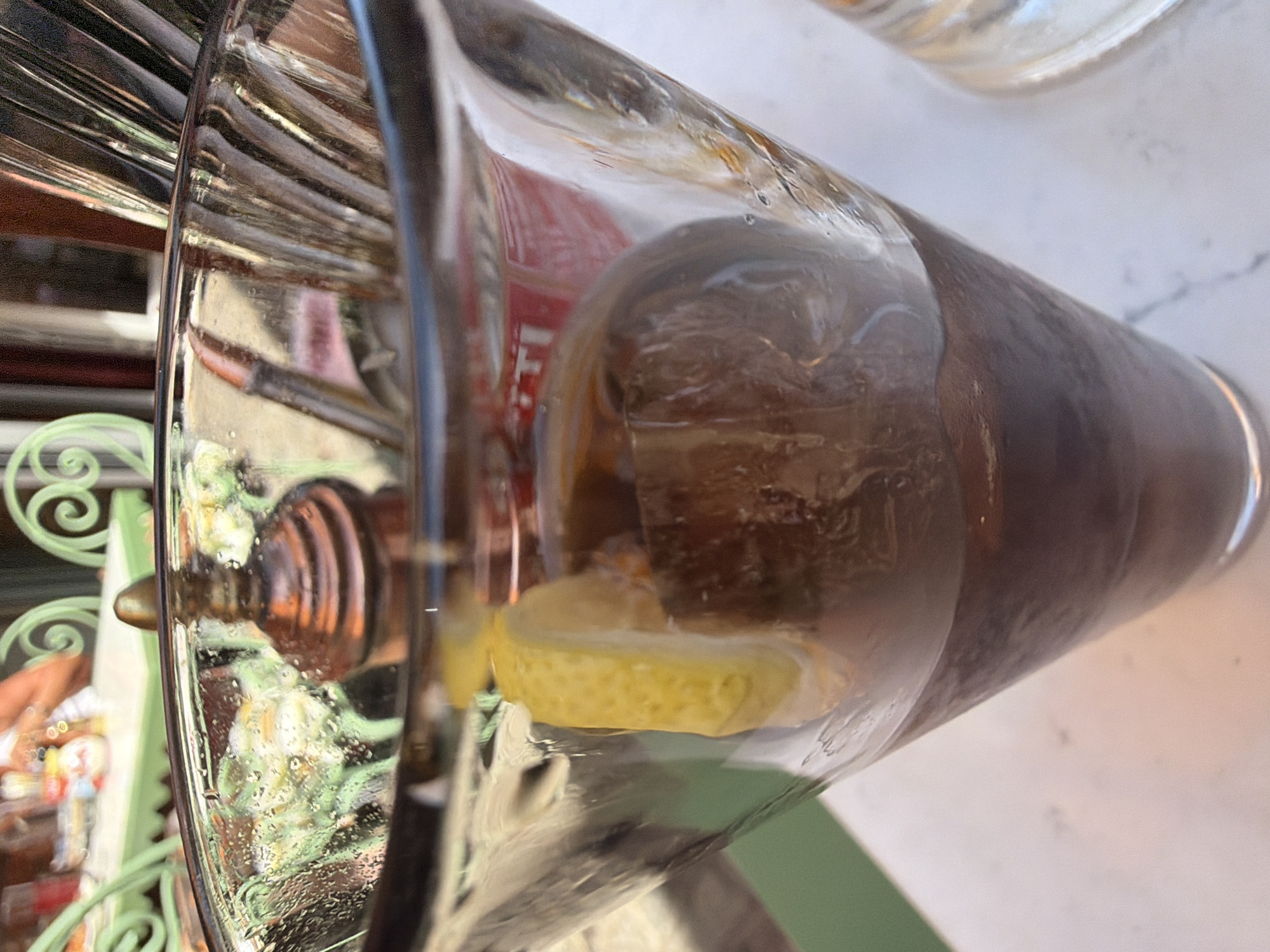 A close-up shot of a glass of iced tea with a lemon wedge shows a condensation-covered glass with a large, spherical ice cube. The beverage is dark brown, suggesting it is iced tea or a similar dark-colored drink. In the background, blurry green chairs and a table with food and drinks can be seen, suggesting an outdoor dining setting. The image does not provide specific details to identify the location in Malmö, Sweden.