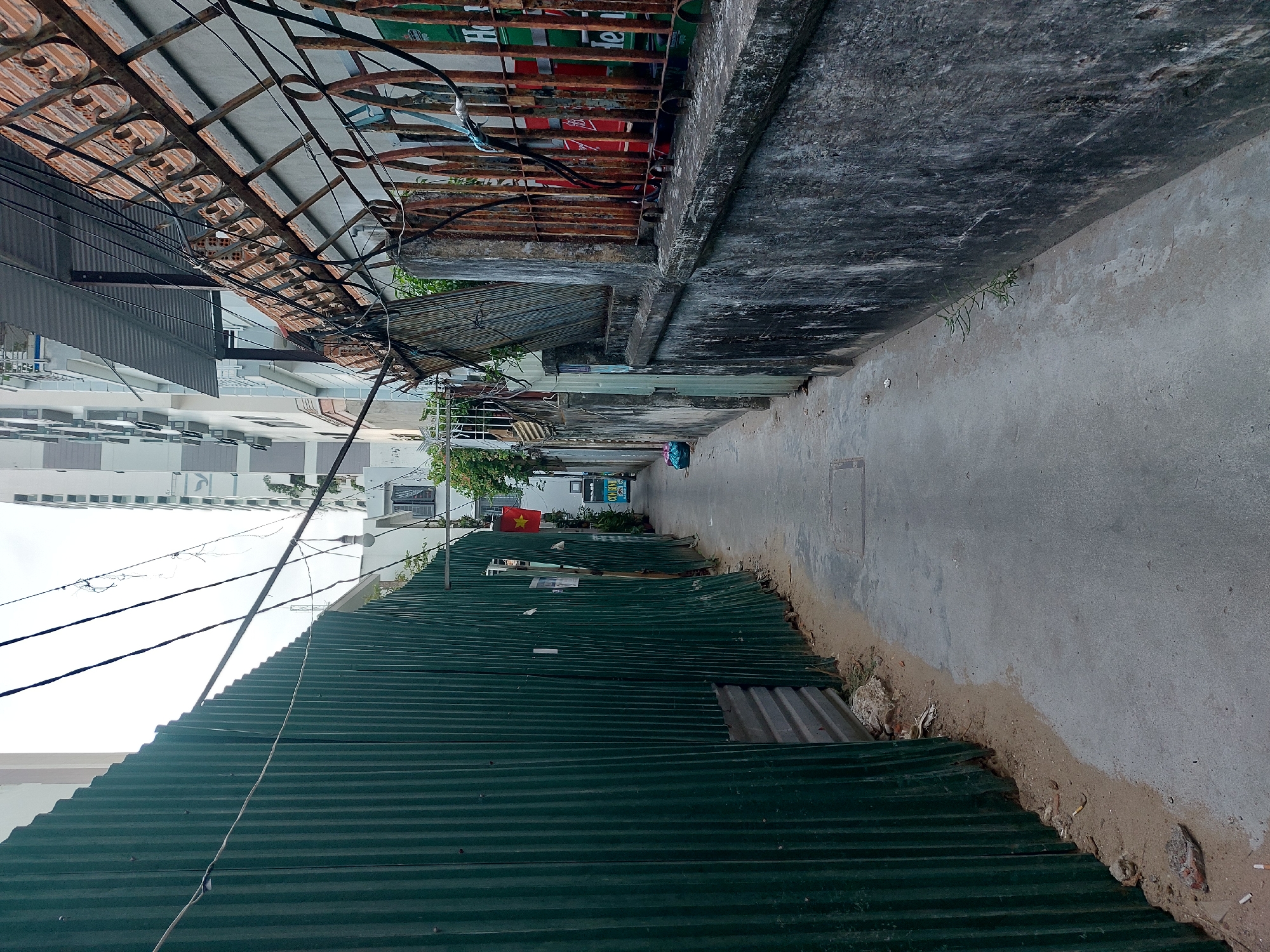 This is a photograph of an alleyway in Nha Trang Ward, Vietnam. The alley is narrow and paved with concrete. On the left side of the alley is a tall, green, corrugated metal fence, which appears to be temporary construction fencing. On the right side of the alley is a concrete wall topped with a rusty metal fence. The metal fence on the right has vertical bars and decorative scrollwork, and behind it, several stacks of what appear to be beer crates or cans are visible, with red and green labels. Electrical wires are strung across the alley overhead. At the far end of the alley, a small Vietnamese flag is visible hanging from a building. A blue plastic bag lies on the ground in the alley. The scene suggests a backstreet or service alley in a densely populated urban area.