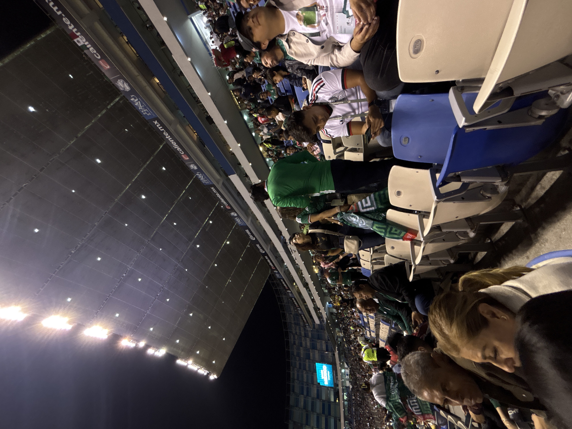 The image shows a stadium filled with people, presumably attending a sporting event. The location is likely in Puebla, Mexico, based on the presence of the Estadio Cuauhtémoc and the "caliente.mx" banners, a common sight in Mexican stadiums. People are seated in stadium chairs, some wearing jerseys of what appears to be the Mexico national team. Some people are looking down at their phones. The stadium lights are on, indicating it is nighttime. The structure of the stadium is visible, including the roof and the seating arrangement.