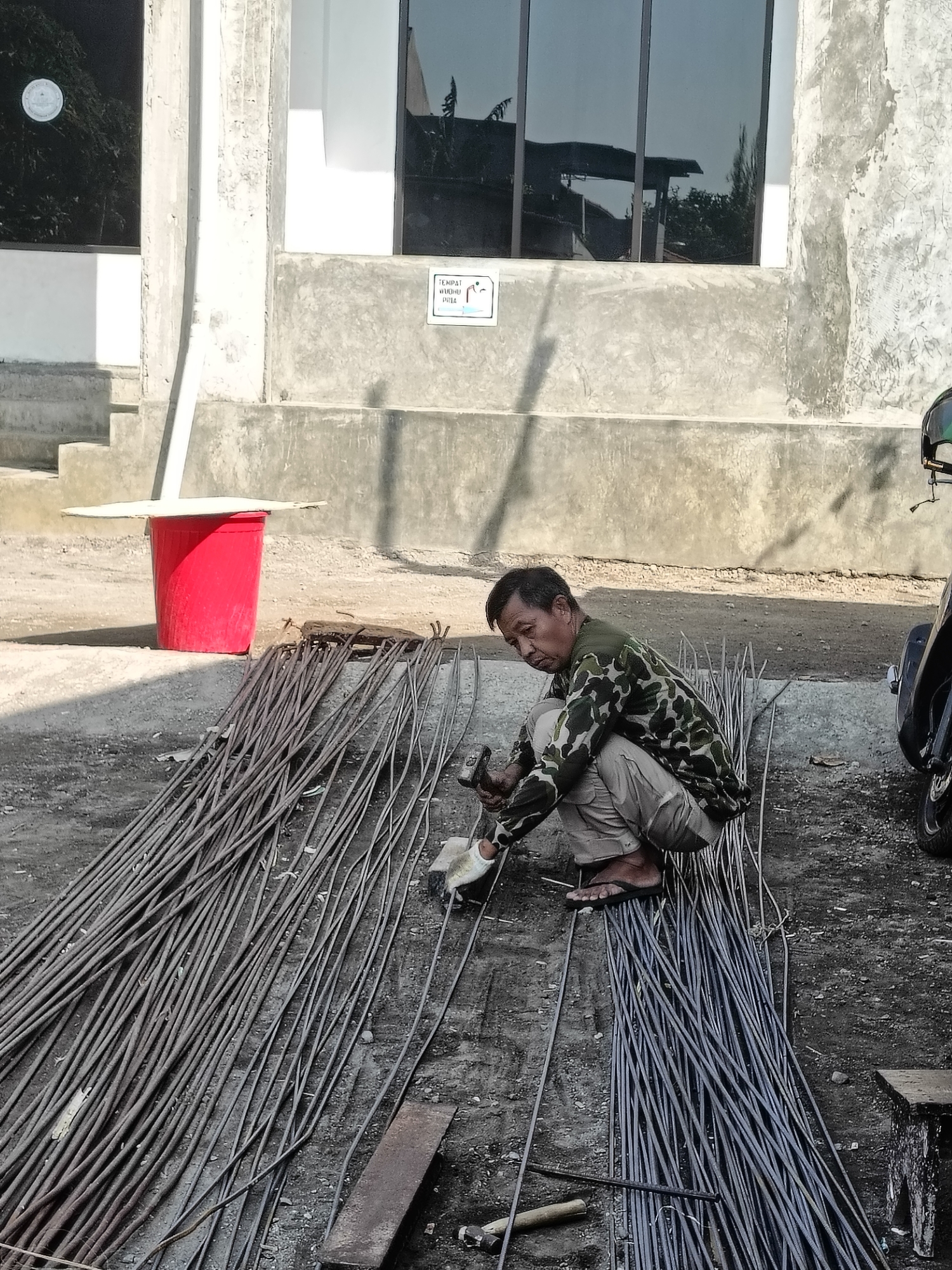 A man is crouched on the ground, working with a pile of rebar. He is wearing a camouflage shirt and light-colored pants. He holds a hammer in his right hand and appears to be working on one of the rebar pieces. To his left, a red bucket sits on a stepped concrete surface. Behind the man, a building with a textured concrete facade and large, reflective windows is visible. A motorcycle is partially visible to the right of the frame. The scene appears to be outdoors, on a paved or dirt surface, with the ground covered in rebar. A sign with Indonesian text is affixed to the wall of the building. The location is Gedangan, Indonesia.