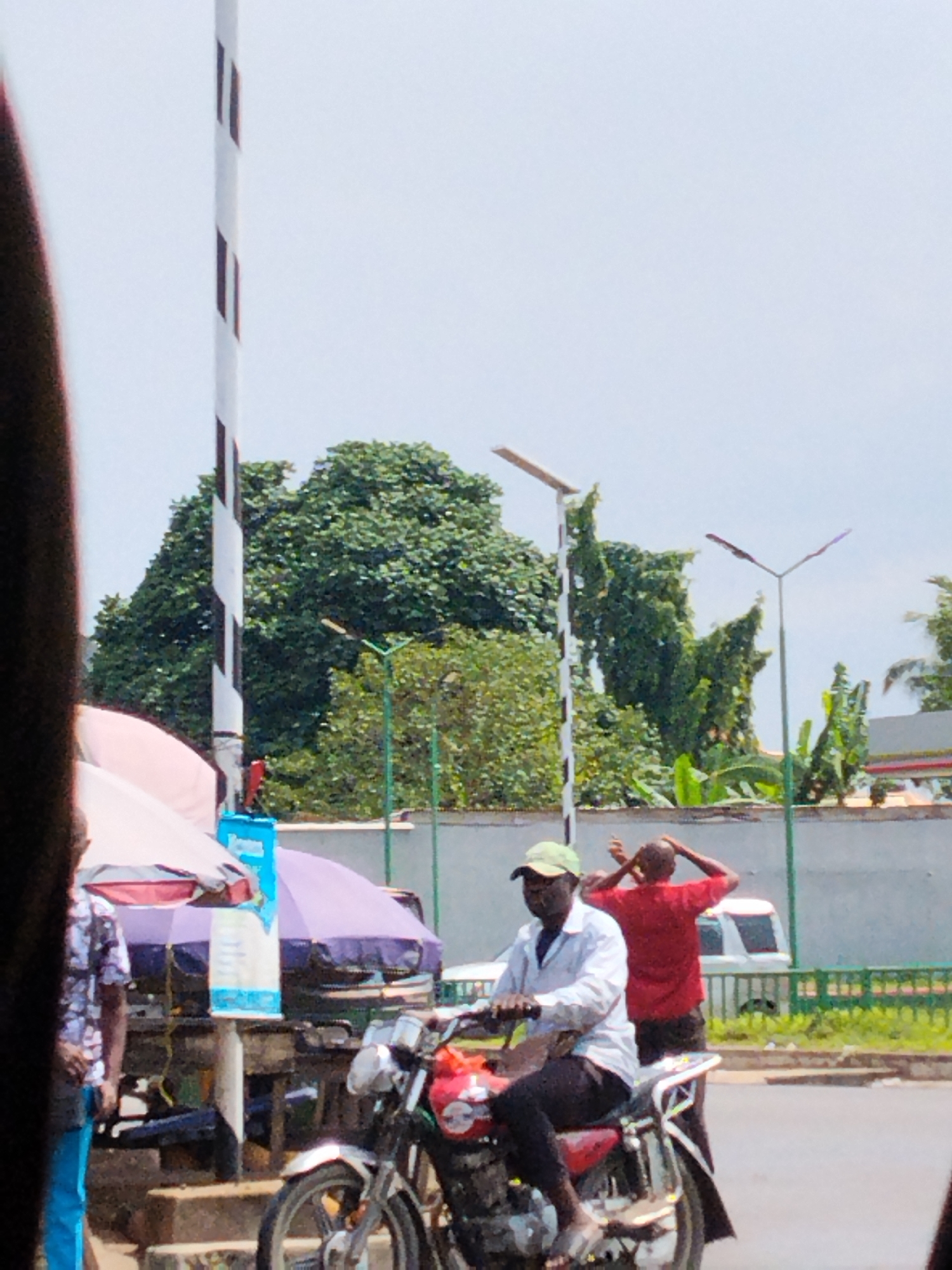 The image captures a street scene in Aba, Nigeria. A man wearing a white shirt and a light green cap is riding a red and silver motorcycle. A man in a red shirt stands nearby with his hands on his head. In the background, there is a white wall with a green fence, and behind it, lush green trees. There's a tall, black and white striped pole adjacent to a roadside stand, which is covered by purple and pink tarpaulins. The sky is overcast. Streetlights are visible in the background.