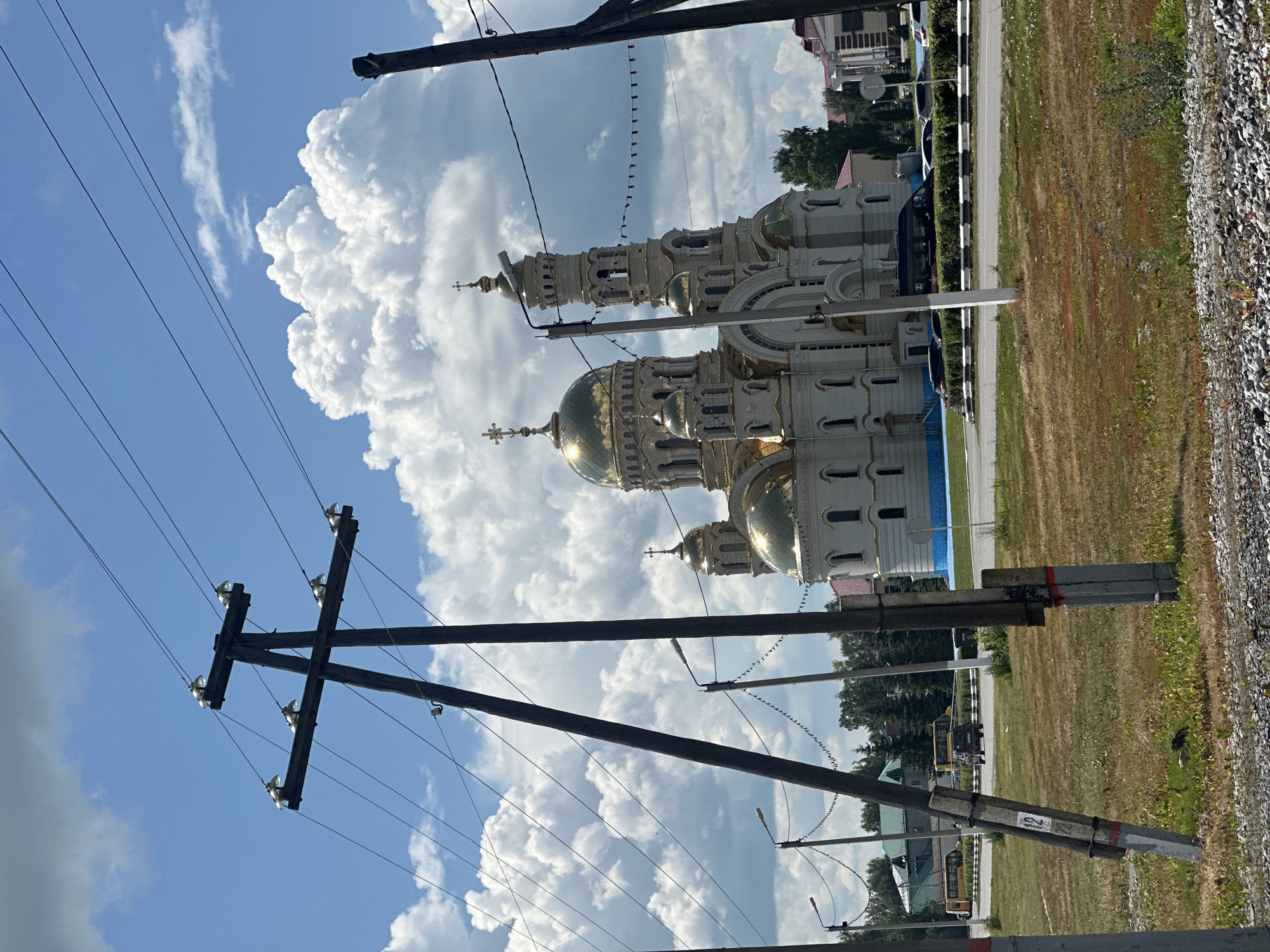 An outdoor, eye-level, medium shot captures a large, ornate church with golden domes against a bright blue sky filled with fluffy white clouds. In the foreground, utility poles with crisscrossing wires dominate the view, partially obscuring the church. A road with some traffic, including a yellow bus and a red vehicle, is visible in front of the church. Trees and buildings are scattered around the church. The ground in the foreground is covered with dry grass and gravel.