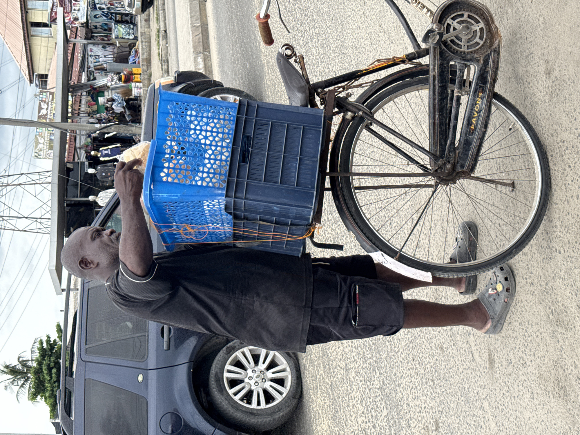 An elderly man with a shaved head stands on a street in Ado, Nigeria, next to a blue SUV and a vintage bicycle marked "AVON" on its back. The man is placing something in a blue plastic crate attached to the bike, positioned in front of the rear wheel. He is wearing a black t-shirt, dark shorts, and gray Crocs. The bicycle is old, with some rust, and features a rear carrier. A building with shops is visible in the background.