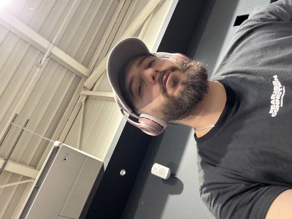 A man wearing a gray cap, headphones, and a black t-shirt with white writing, looks directly at the camera. He has a thick, dark beard and a silver chain around his neck. He is in a room with a corrugated metal ceiling and a large, white unit on the wall. A black wall is also visible, with a small, white object attached to it. The camera angle is low, tilting upwards.