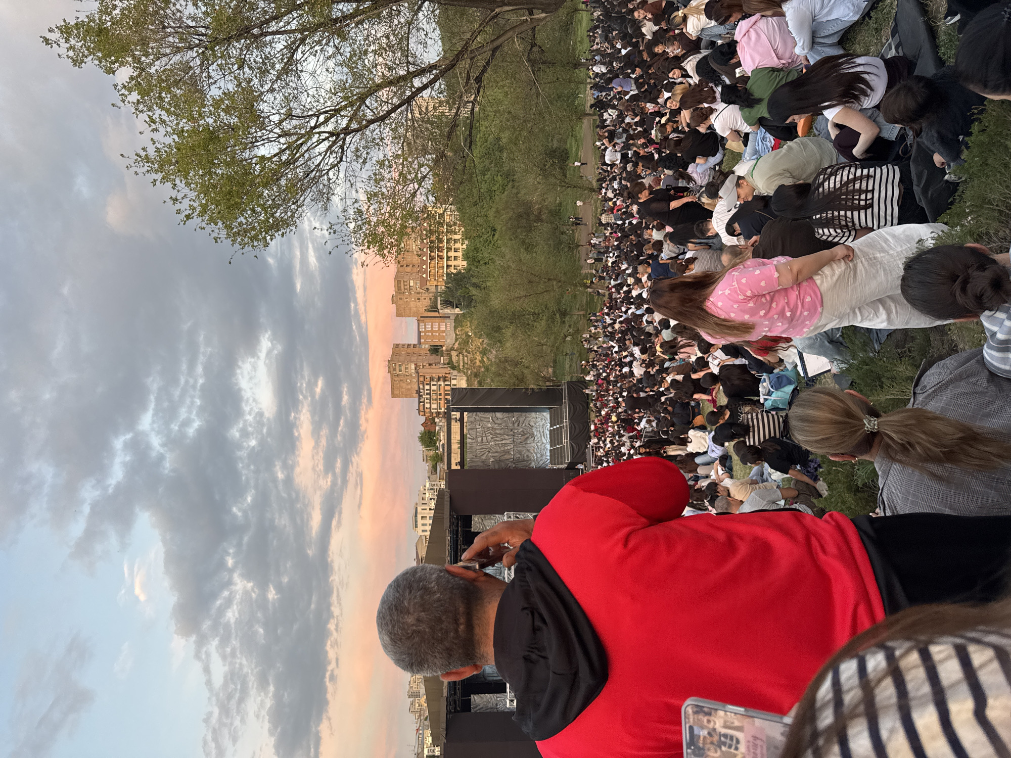 This image depicts an outdoor evening event in Yerevan, Armenia, at dusk. A large crowd of people is gathered on a grassy, gently sloping terrain, facing a substantial performance stage.
In the immediate foreground, a man with short grey hair, wearing a red long-sleeved top with a black hoodie, is seen from behind, holding a dark-colored smartphone to his right ear. To his left, the edge of another smartphone screen is partially visible, displaying an image. To the man's right, a woman in a light pink shirt adorned with white hearts and loose-fitting white pants stands, facing away from the camera towards the stage. Further right and slightly lower, another woman with light brown hair tied in a ponytail, wearing a grey plaid jacket, is seated, also facing the stage.
The midground is dominated by a dense audience, comprising hundreds of individuals, mostly seated on the grass or on blankets, facing the stage. The majority of the attendees appear to be young adults. The stage itself is a large, dark, rectangular structure with two prominent vertical panels on either side of a central opening, featuring a textured or abstract pattern resembling crumpled material. Above the stage, a dark canopy or roof extends.
Behind the stage and the immediate audience, a line of deciduous trees with green foliage marks the edge of the park area. Beyond these trees, on an elevated landscape, numerous multi-story residential buildings, likely apartment blocks characteristic of urban architecture, are visible under the twilight sky.
The sky in the background displays a gradient of colors, with soft pink and orange hues near the horizon, transitioning to light blue and grey further up, interspersed with scattered clouds. A large tree with visible branches and green leaves is present on the right side of the image, partially obscuring the view of the sky and some distant buildings. The lighting suggests late afternoon or early evening, with the sun setting, casting a warm glow on the clouds.