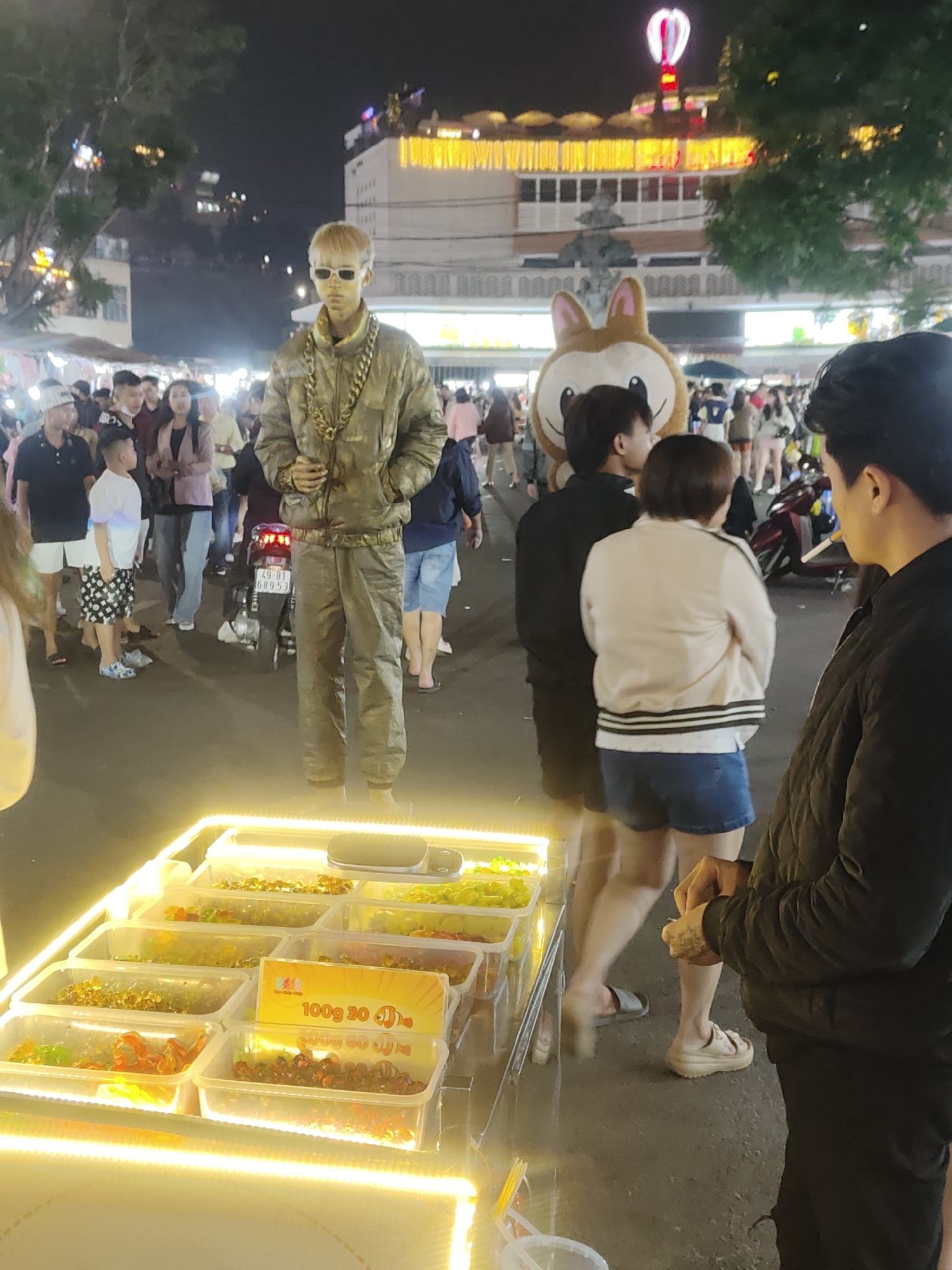 In Da Lat, Vietnam, an outdoor market at night is illuminated by bright lights. A man dressed as a statue, covered in gold paint and wearing oversized chains, stands motionless. He is in front of a brightly lit display of candies and snacks. Behind him, a crowd of people mill about, some looking at the display, others talking. A person in a large animal costume stands near the statue. In the background, buildings are visible, with lights and signs indicating the lively atmosphere of the city. A motorcycle is parked nearby.