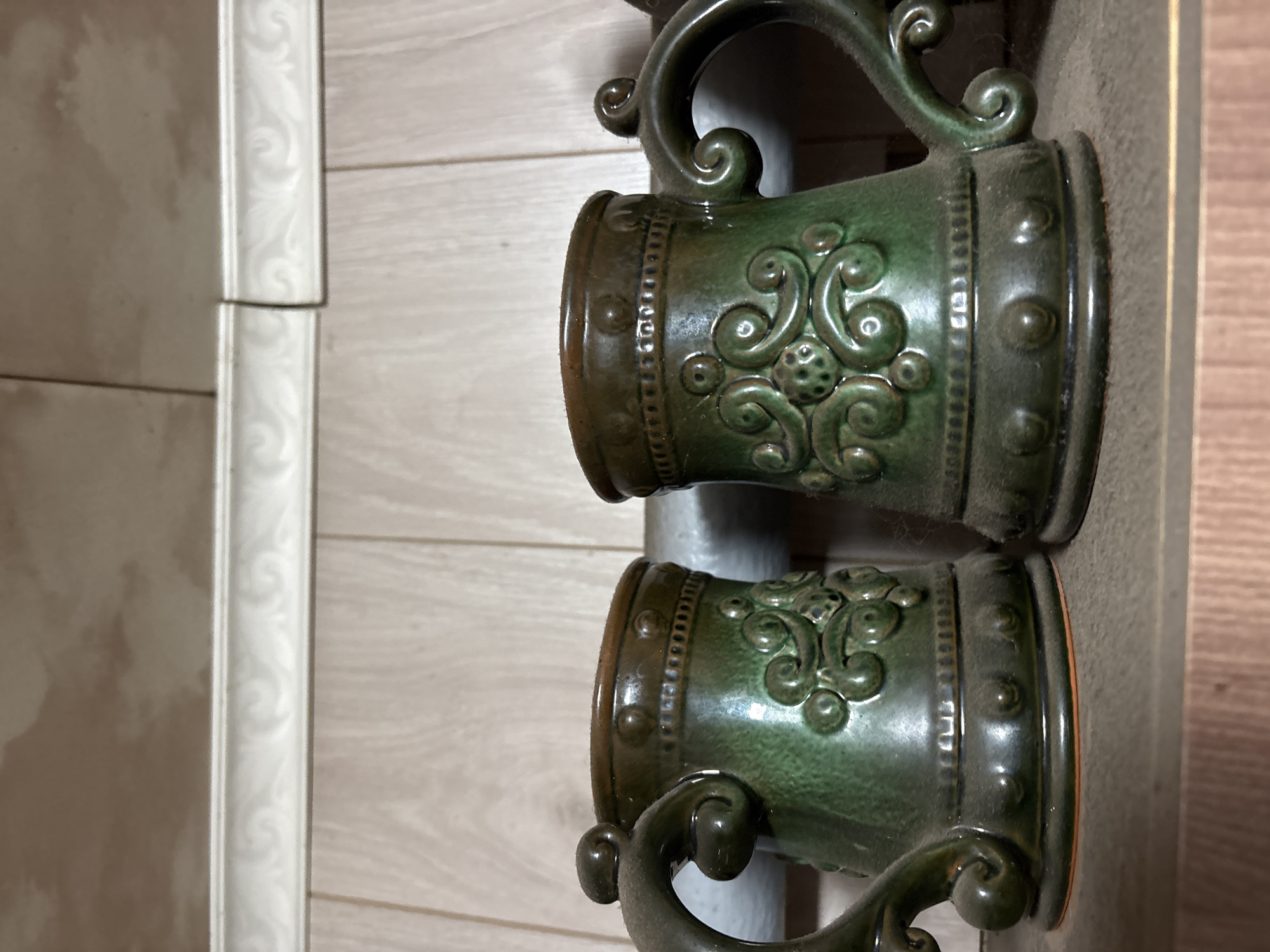 The image shows a close-up of two green ceramic mugs with ornate designs, possibly on a shelf. The mugs are positioned side-by-side, with the left mug slightly closer to the viewer. Both mugs have handles with decorative swirls and a band of raised dots around the rim. The body of each mug features a raised, intricate pattern of swirls and small spheres. The mugs appear dusty, suggesting they may be old or infrequently used. The background consists of a light-colored, wood-paneled wall with a decorative white trim running horizontally across it. The lighting is diffuse, casting soft shadows. The image was taken in the city of Vetluzhsky, Russia.