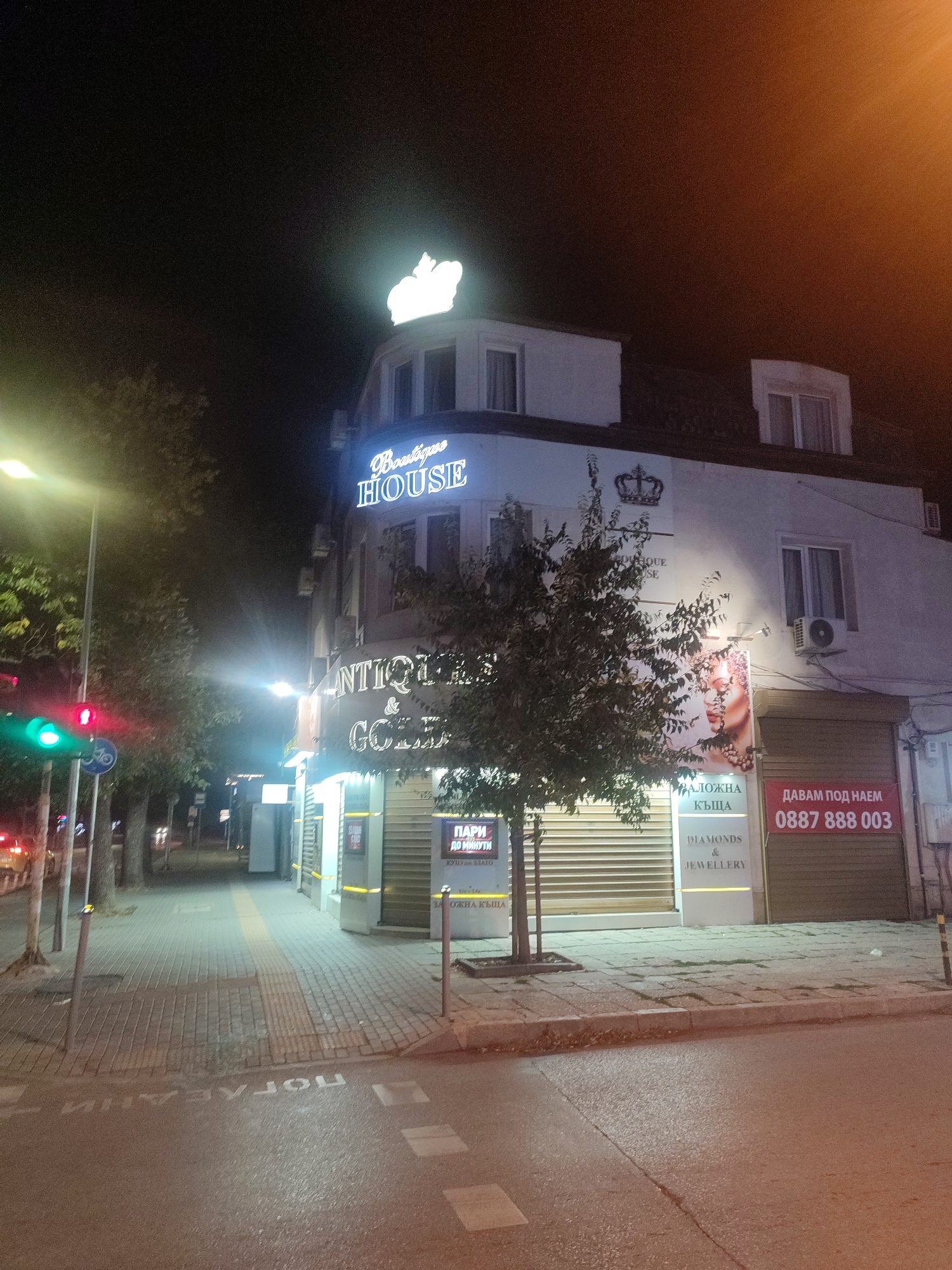 The image depicts a nighttime scene on a street in Varna, Bulgaria, specifically on or near General Skobelev Street. A multistory building with a white facade is illuminated by artificial lights. A prominent neon sign reads "Boutique HOUSE" with a crown emblem above it. Below this, another sign indicates "ANTIQUES & GOLD". Further down the building, a large advertisement poster features a woman's face. Adjacent to this, there are signs for "DIAMONDS & JEWELLERY" and a red banner stating "ДАВАМ ПОД НАЕМ 0887 888 003" (which translates to "FOR RENT").
To the left of the building, a sidewalk is visible, lined with trees. A traffic light shows a red and green signal. The street itself is paved, and in the foreground, white markings on the asphalt are visible, possibly indicating a pedestrian crossing or road markings. The overall environment is that of a commercial street at night, with businesses and illuminated signage. There are no people clearly visible in the image. The lighting suggests it is late evening or night.