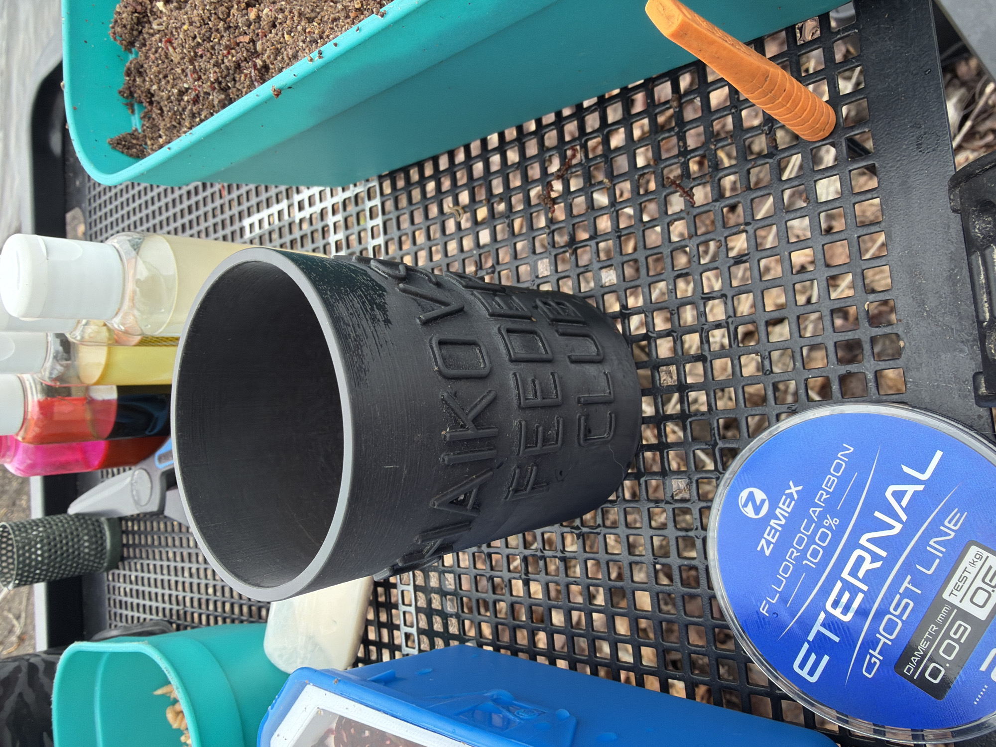 The image is a close-up shot of various fishing-related items arranged on a black, grid-like surface, likely a fishing platform or table.
Objects:
- A black cylindrical container with raised lettering (possibly "ZEMEX" or a similar brand).
- A spool of blue Zemex "Eternal" fluorocarbon ghost line, labeled as 100% fluorocarbon, with a diameter of 0.09mm and a test strength of 0.5kg.
- Several small bottles with white caps and varying colored liquids, possibly scents or additives.
- A green mesh-like container.
- Other blue and turquoise containers, with fishing bait inside.
- An orange plastic item.
Location:
While the image itself does not provide enough specific details to confirm the location as Chaikovsky, Russia, the presence of the "ZEMEX" brand of fishing line suggests it's possible, as Zemex is a brand that originates in that region.
Description:
The scene is organized, showing the items possibly laid out for fishing.