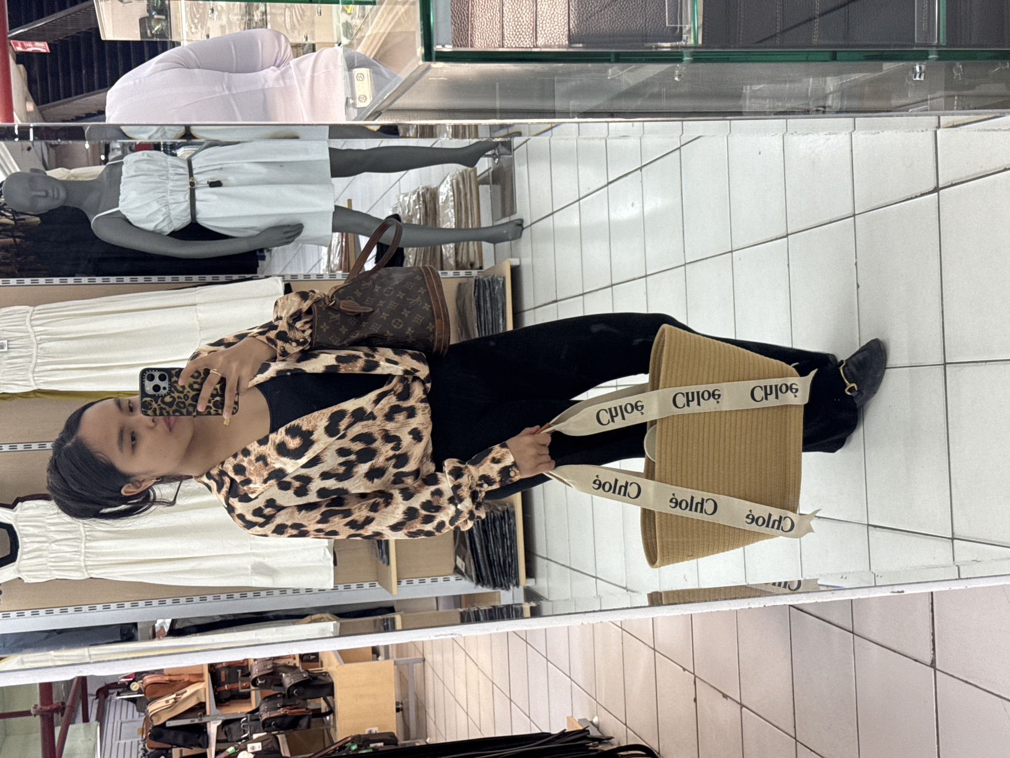 A young woman stands in front of a floor-to-ceiling mirror, taking a selfie with her smartphone. She is wearing a leopard print blouse, black pants, and black loafers. She is holding a straw tote bag with white straps that say "CHLOÉ" in black letters. In the background, the mirror reflects mannequins wearing dresses, racks of clothing, and shelves filled with bags. The floor is tiled. The scene appears to be inside a clothing store, possibly in Davao City, Philippines, given the reference in the prompt.