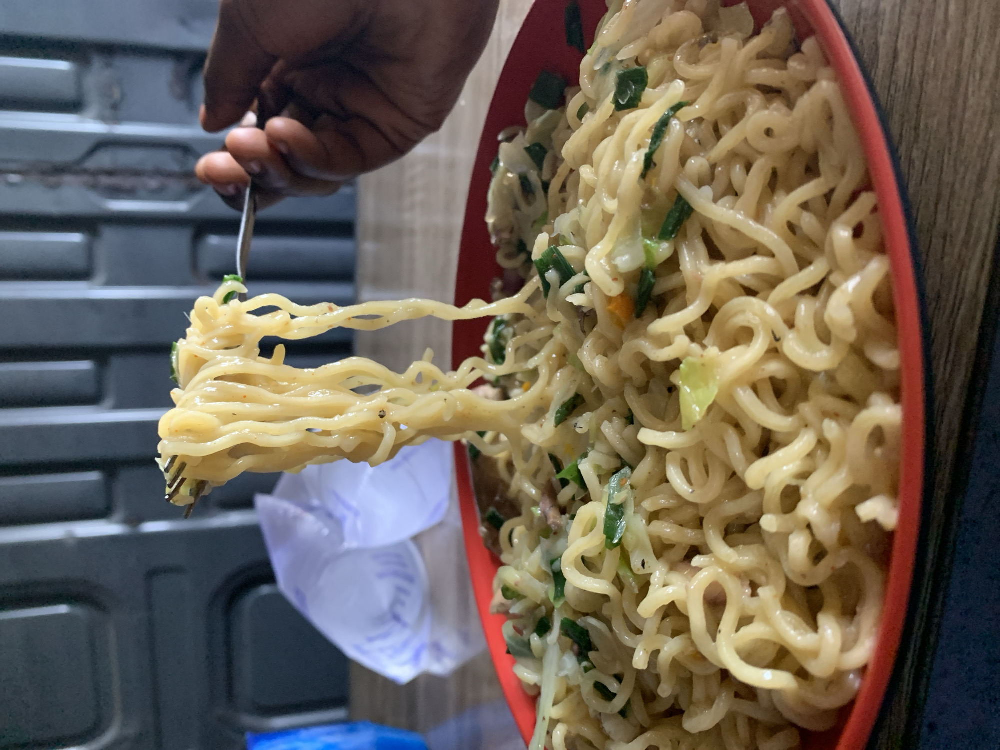 A person's hand is holding a fork laden with noodles over a red bowl filled with more noodles. The noodles appear to be of the instant ramen variety, possibly stir-fried with vegetables like green onions and cabbage. The background is somewhat blurred, but suggests a room with a blue container and plastic bag. The photograph was most likely taken indoors. There is nothing in the image that indicates Rafin Par, Nigeria.
